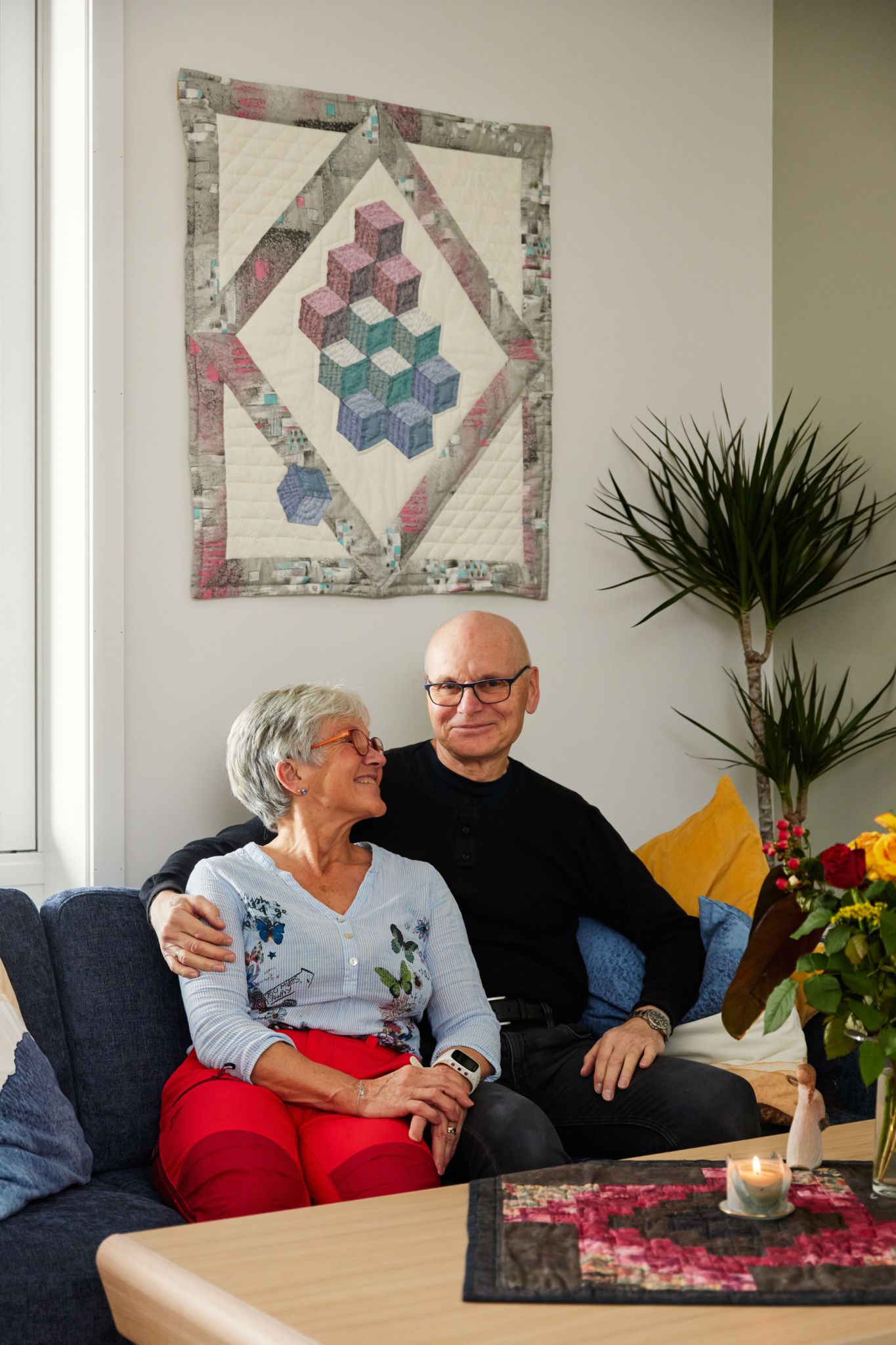 Foto av Solveig og Per i stua si, foran et quilted teppe på veggen