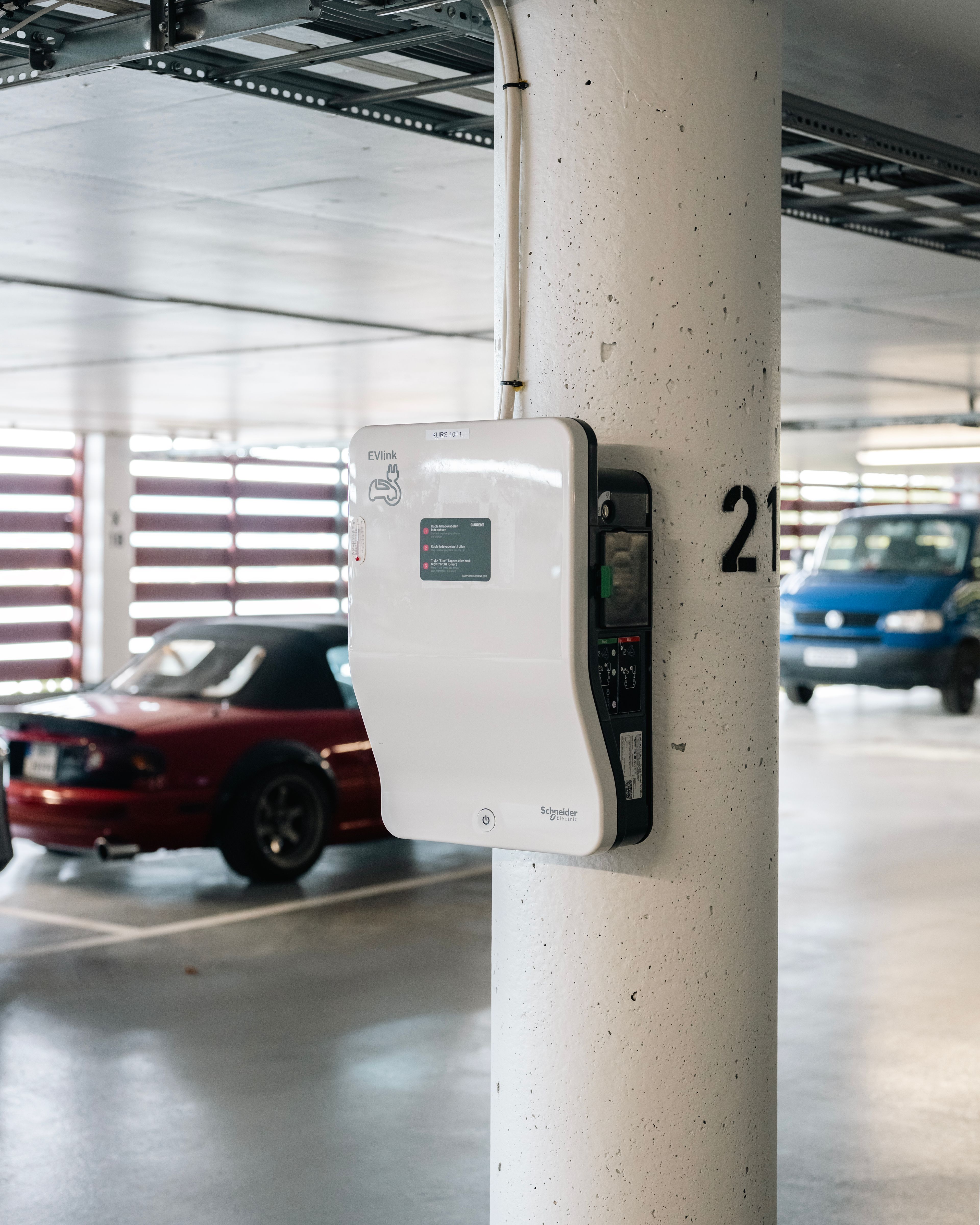 Ladeboks henger på en pullert i parkeringshuset. En rød bil synes i bildet.