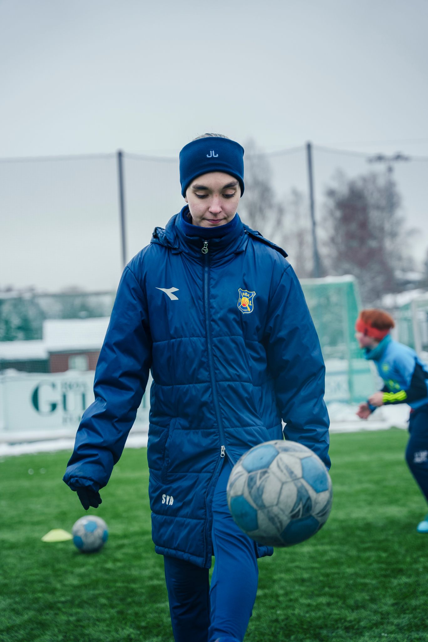 Bilde av Sara som trikser med en fotball ute på banen. Hun har på seg helblå jakke og bukse med logen til Røa.