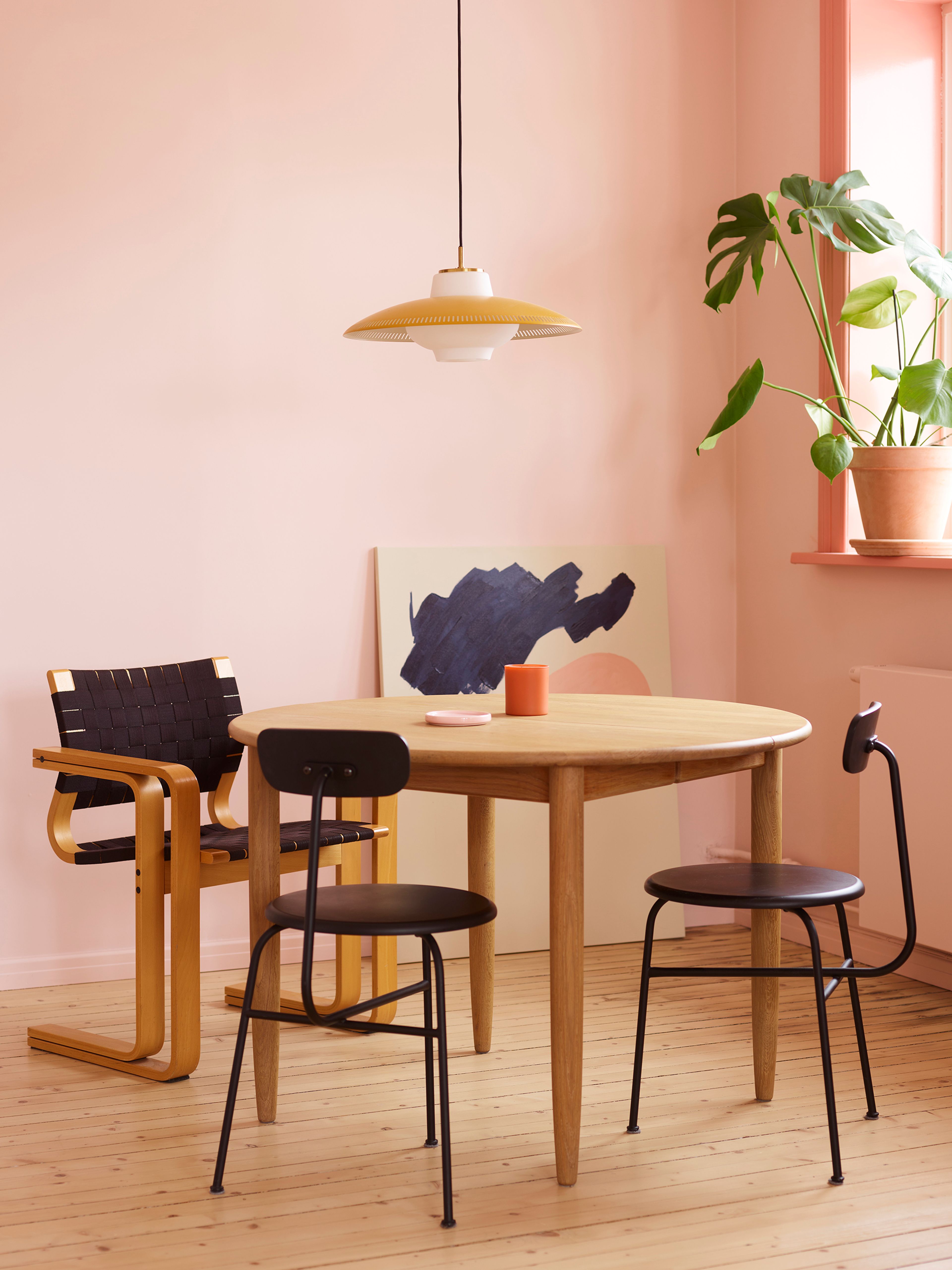 Rundt spisebord i tre med tre svarte stoler og en gul lampe i et rosa rom.