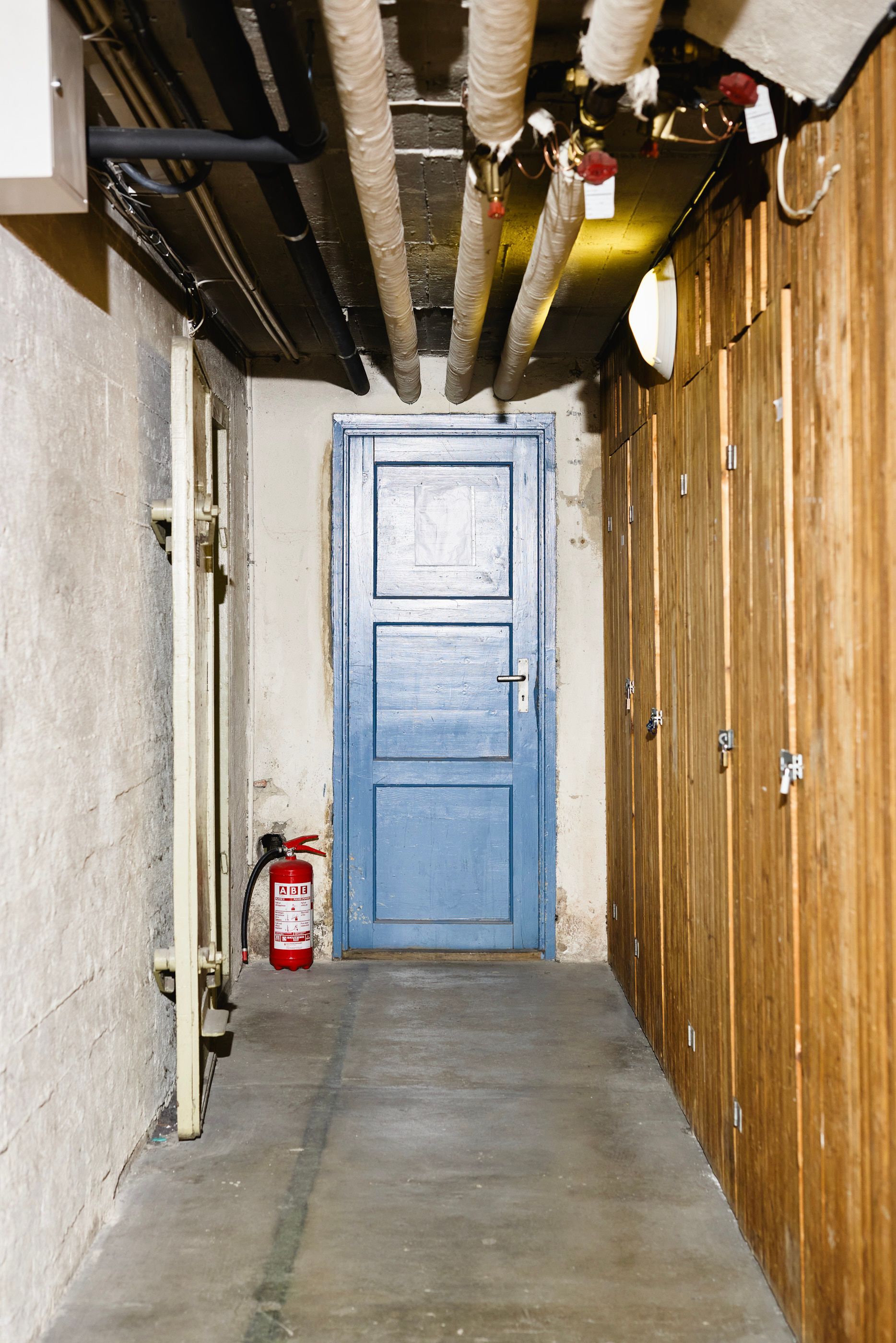 Kjellergang med rør i taket, en blå dør i enden, brannslukningsapparat på gulvet og boder med låsbare tredører langs veggen.
