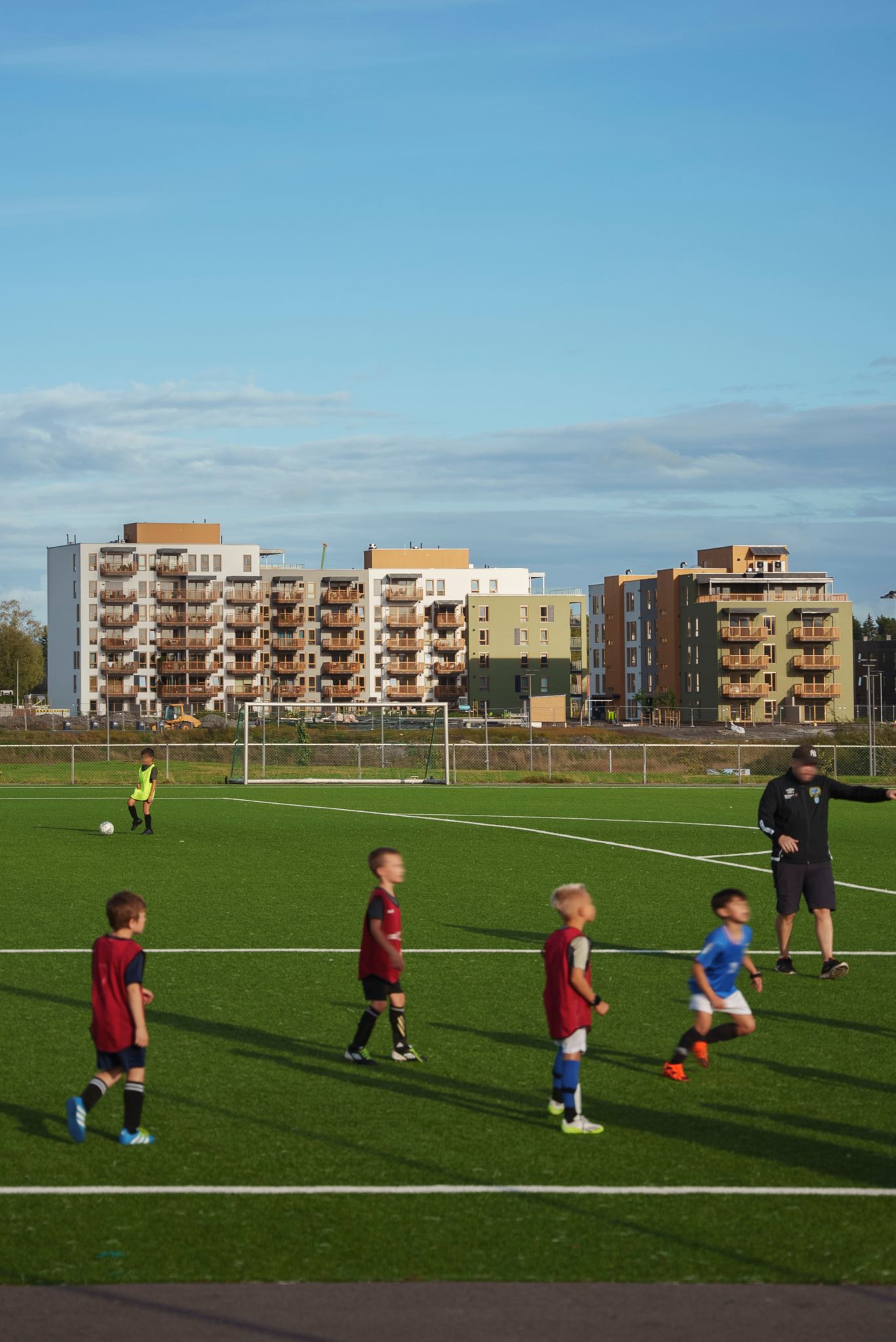 En fotballbane i solen med noen barn som løper, i bakgrunnen synes blokkene i Asper Hageby