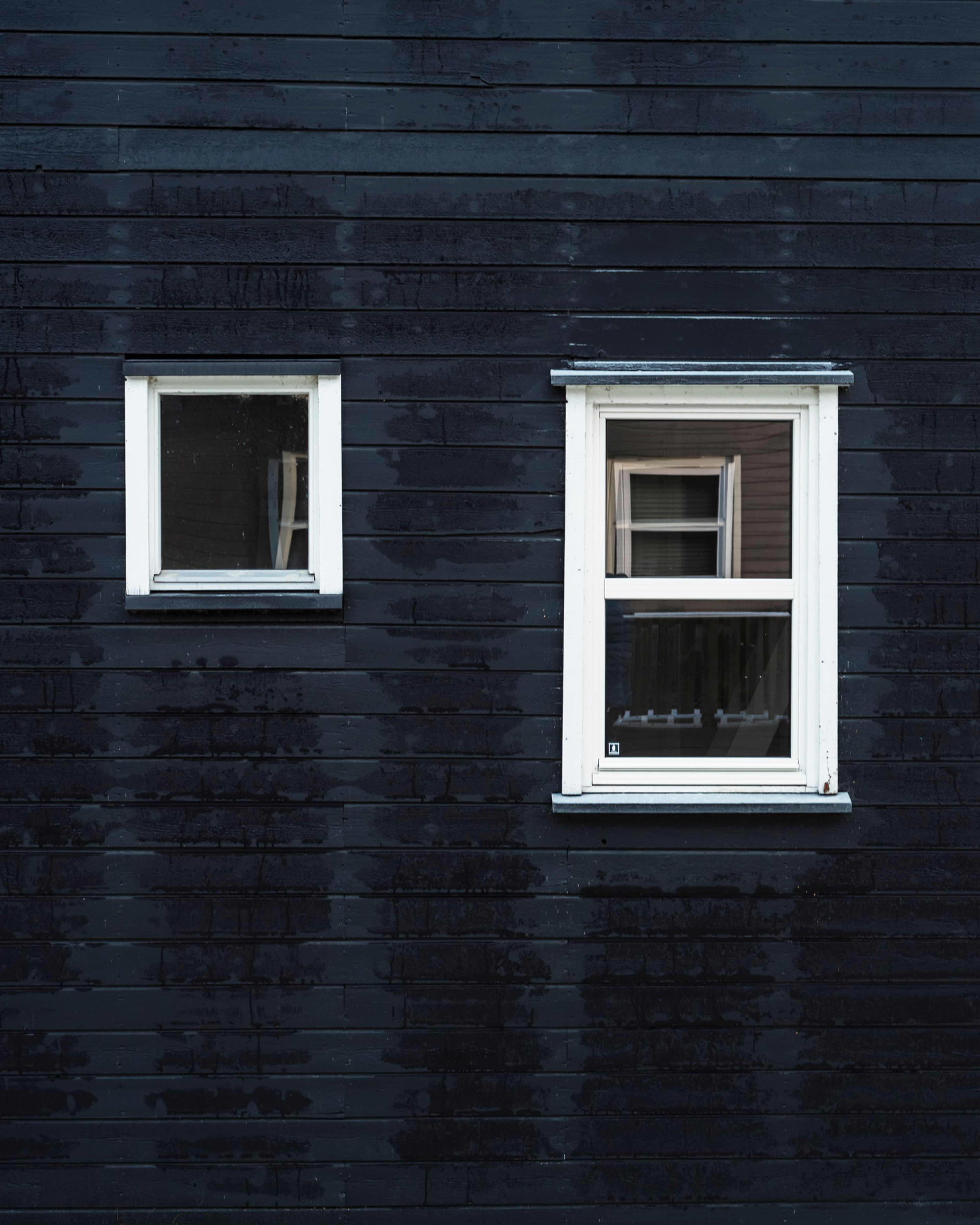 Yttervegg på fritidsbolig med to vinduer og synlig slitasje på kledningen rundt.