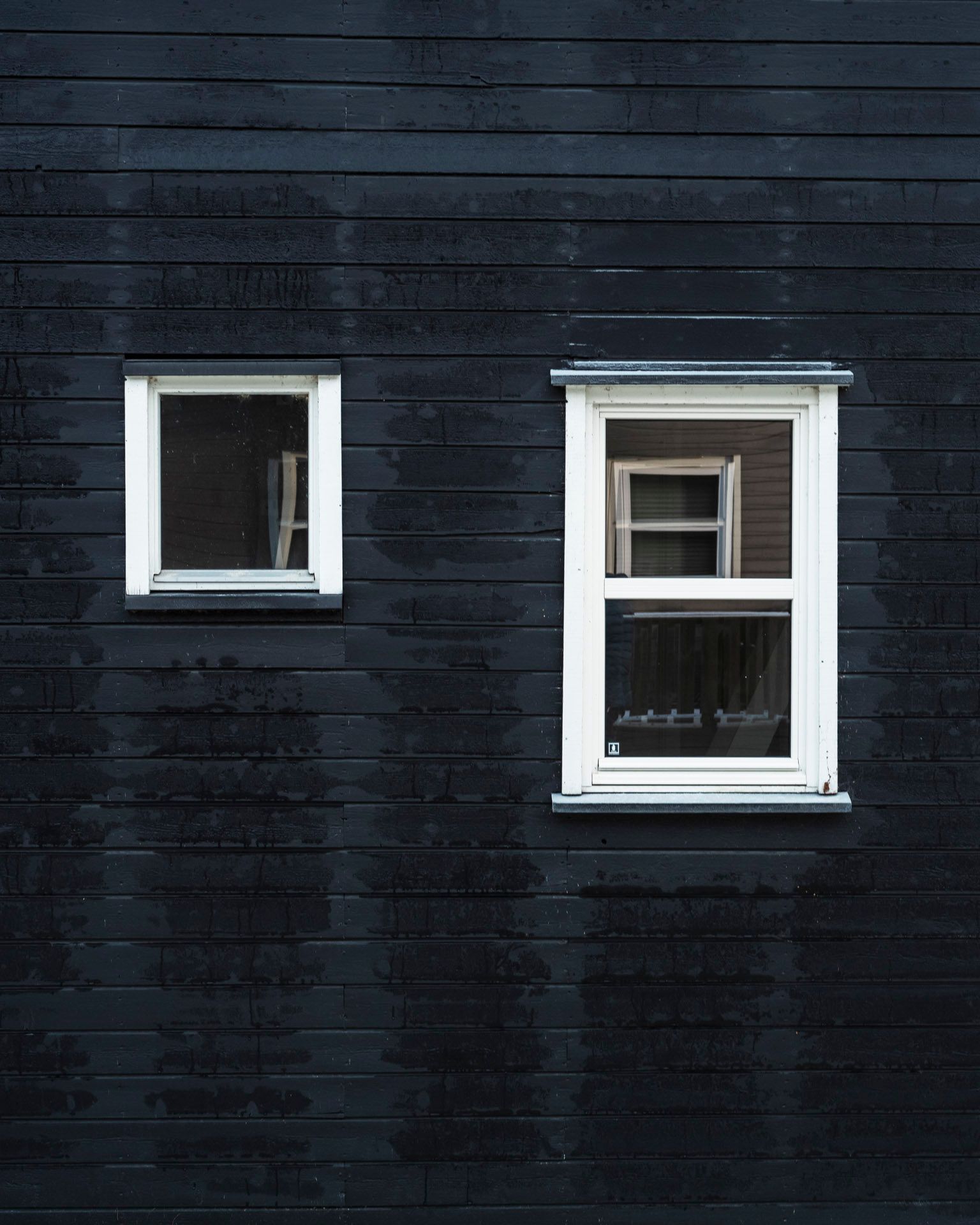 Yttervegg på fritidsbolig med to vinduer og synlig slitasje på kledningen rundt.