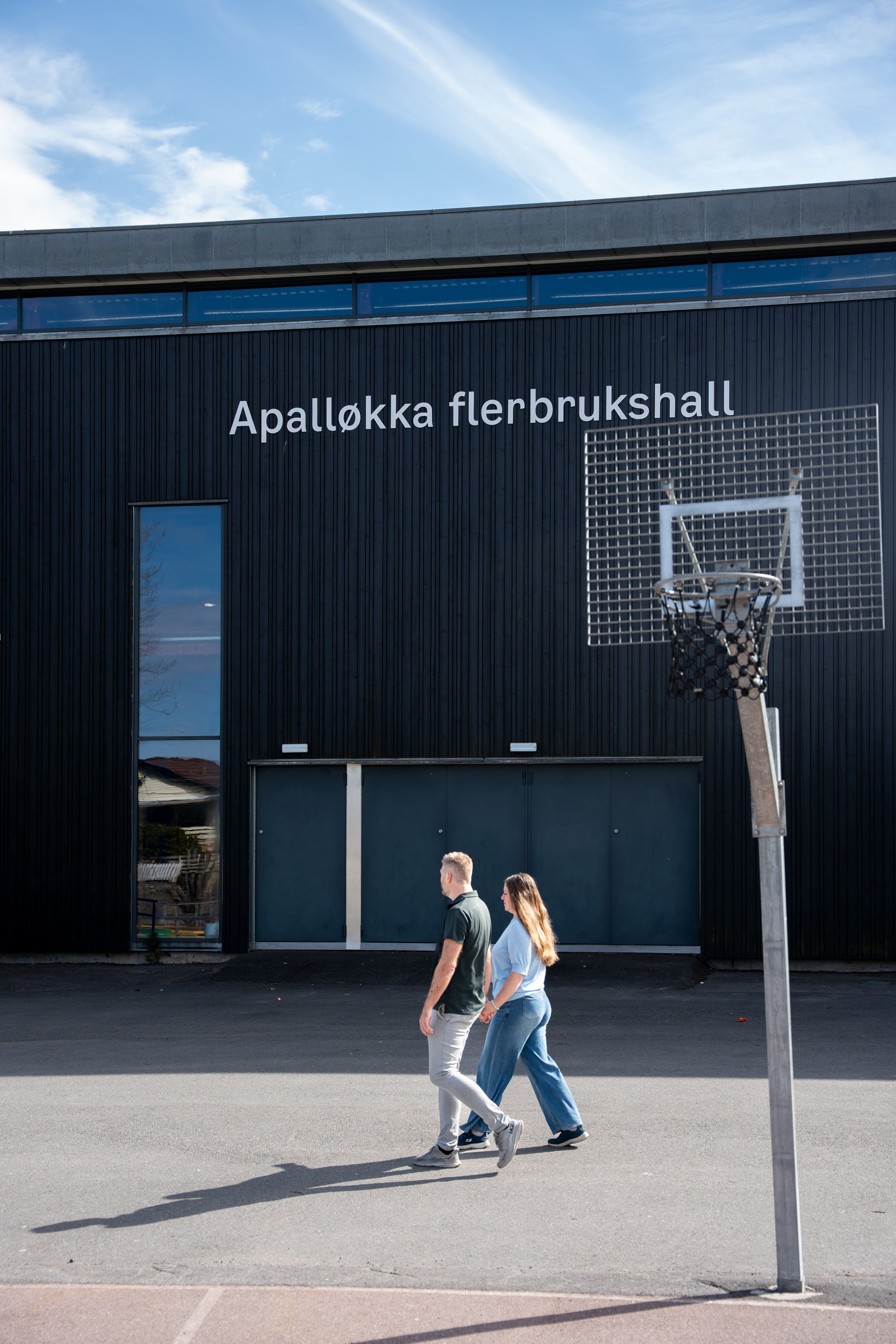 Et par går hånd i hånd foran en mørk bygning med teksten «Apalløkka flerbrukshall» og en basketballkurv.
