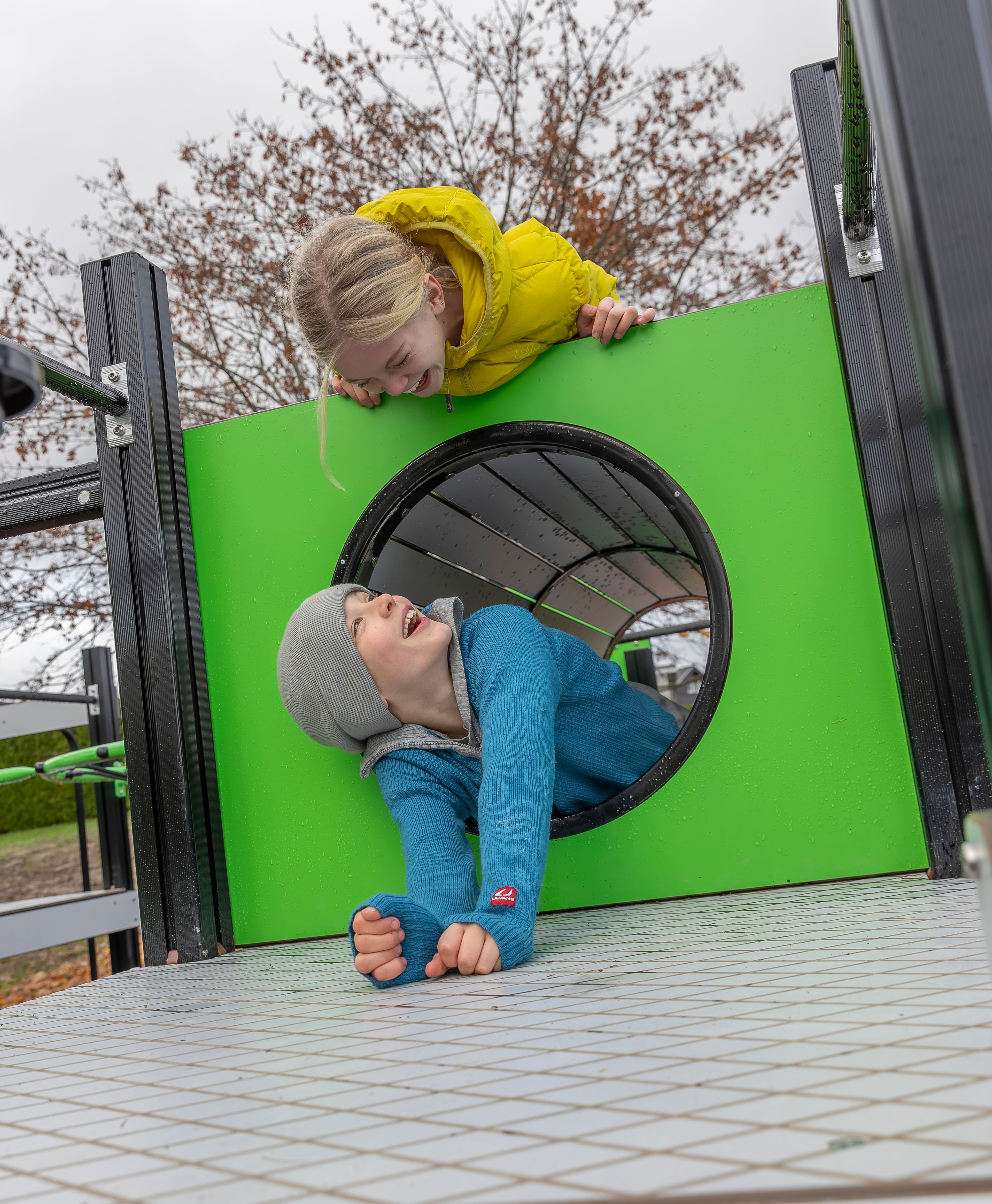 To barn leker sammen i et grønt lekeapparat, der den ene titter ned gjennom et hull mens den andre kryper ut av tunnelen.