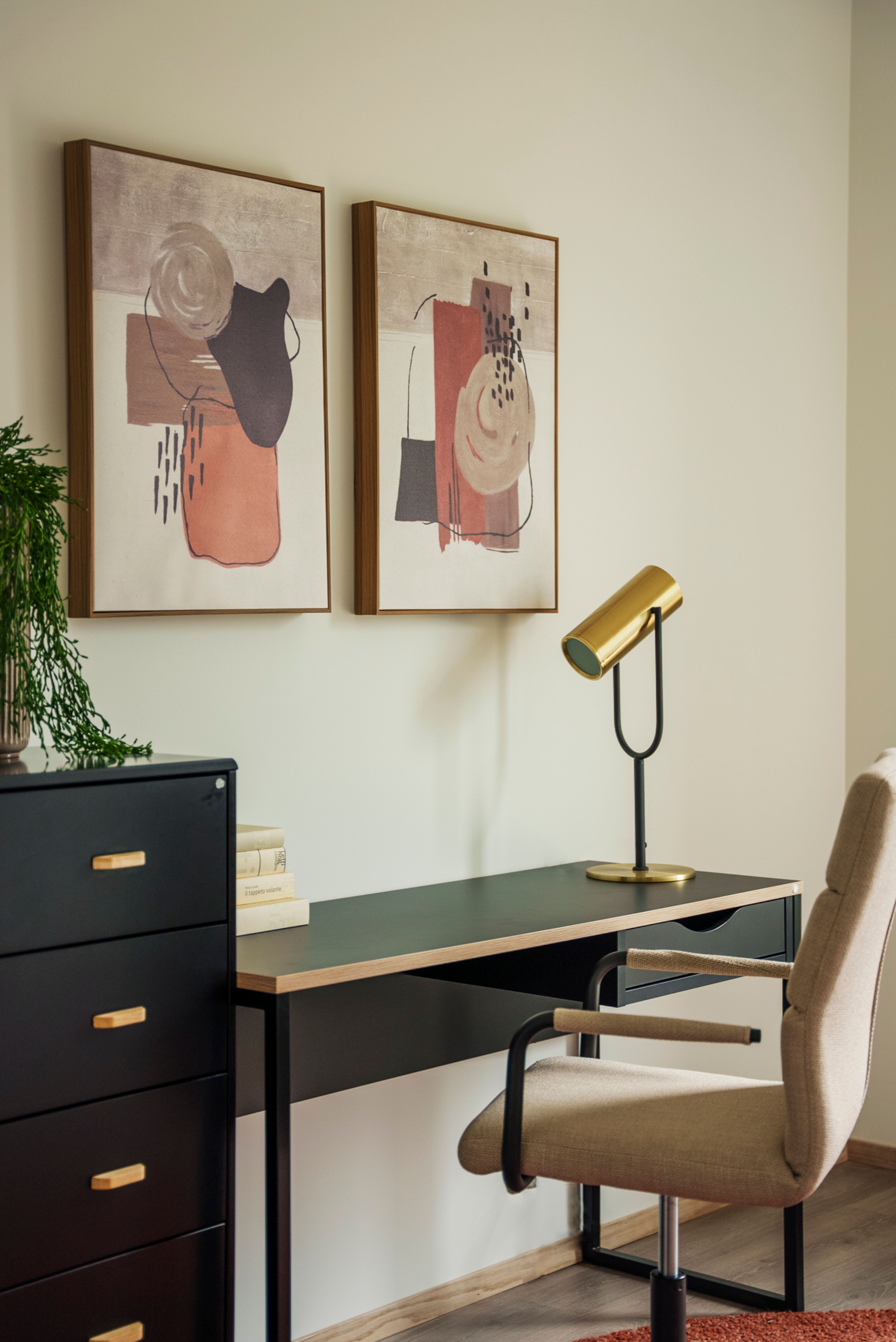 En kontorplass med lampe i gull, beige stol og et arkivskap i sort, dekorert med to malerier med rosa detaljer.