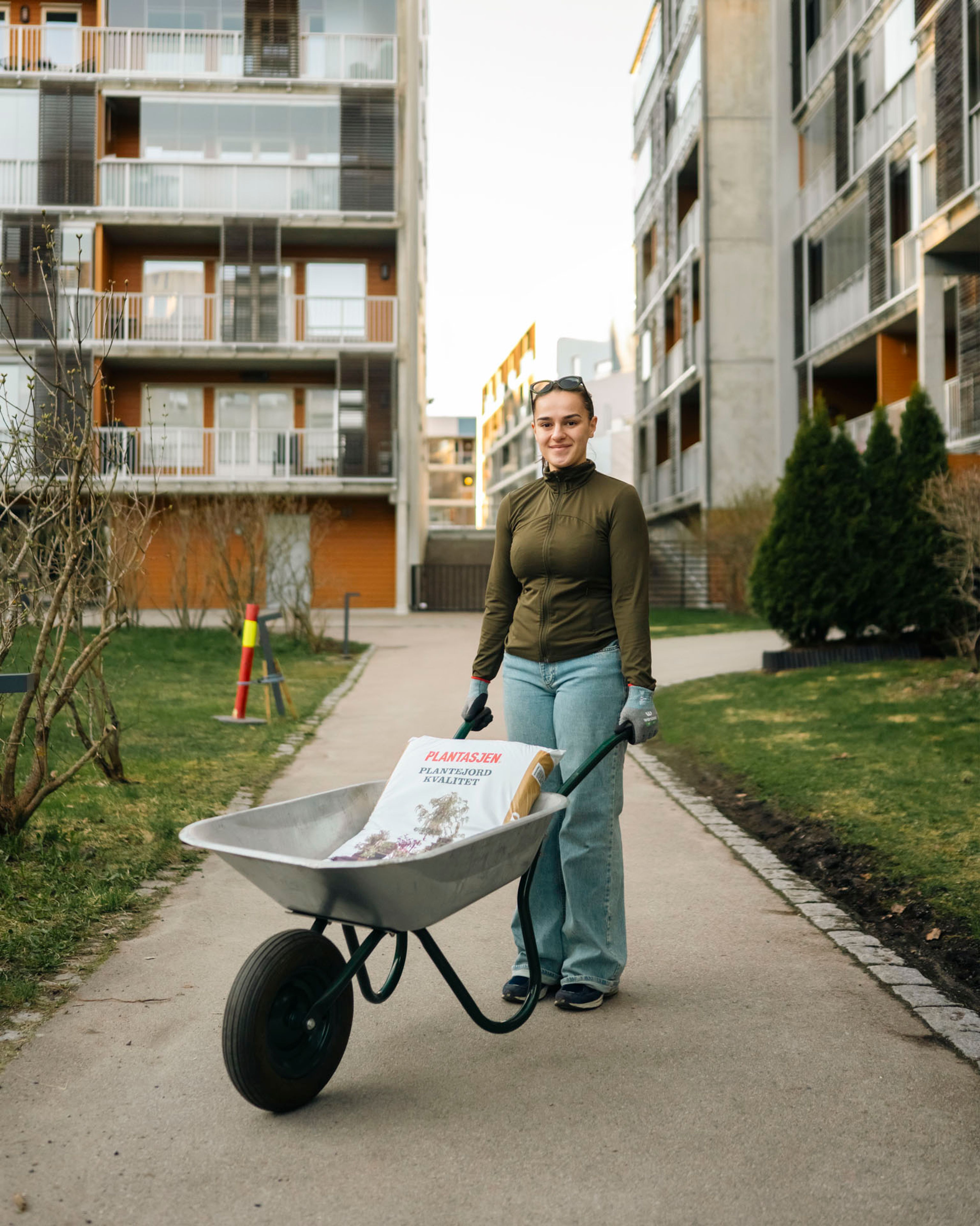 Ung kvinne triller en trillebår med en sekk plantejord på en gangvei mellom to boligblokker.