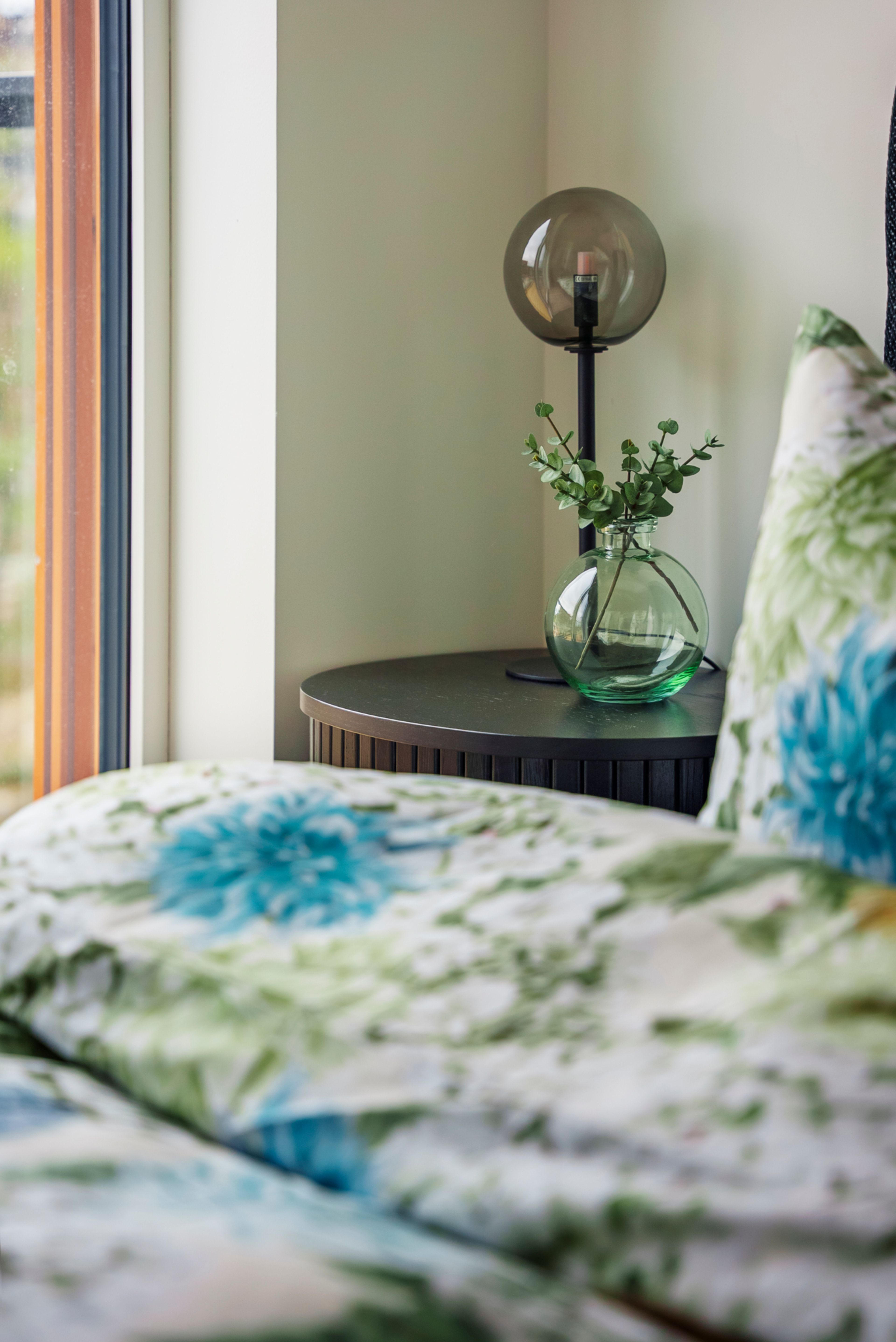 Nærbilde av et blomstrete sengetøy med sort nattbord og lampe i bakgrunnen.