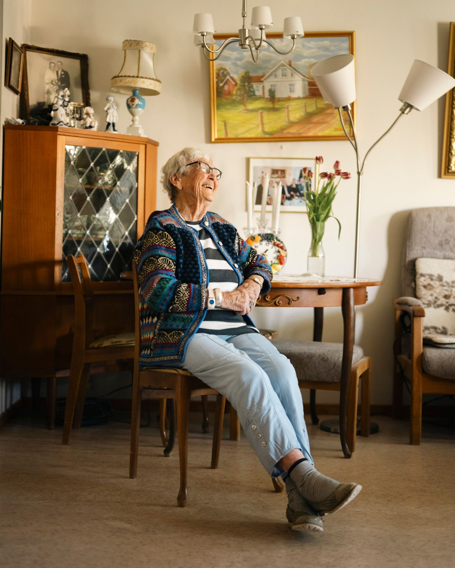 Randi Karlsen (95) sitter smilende i stua si i stripete genser og mønstret jakke, omgitt av malerier og blomster.