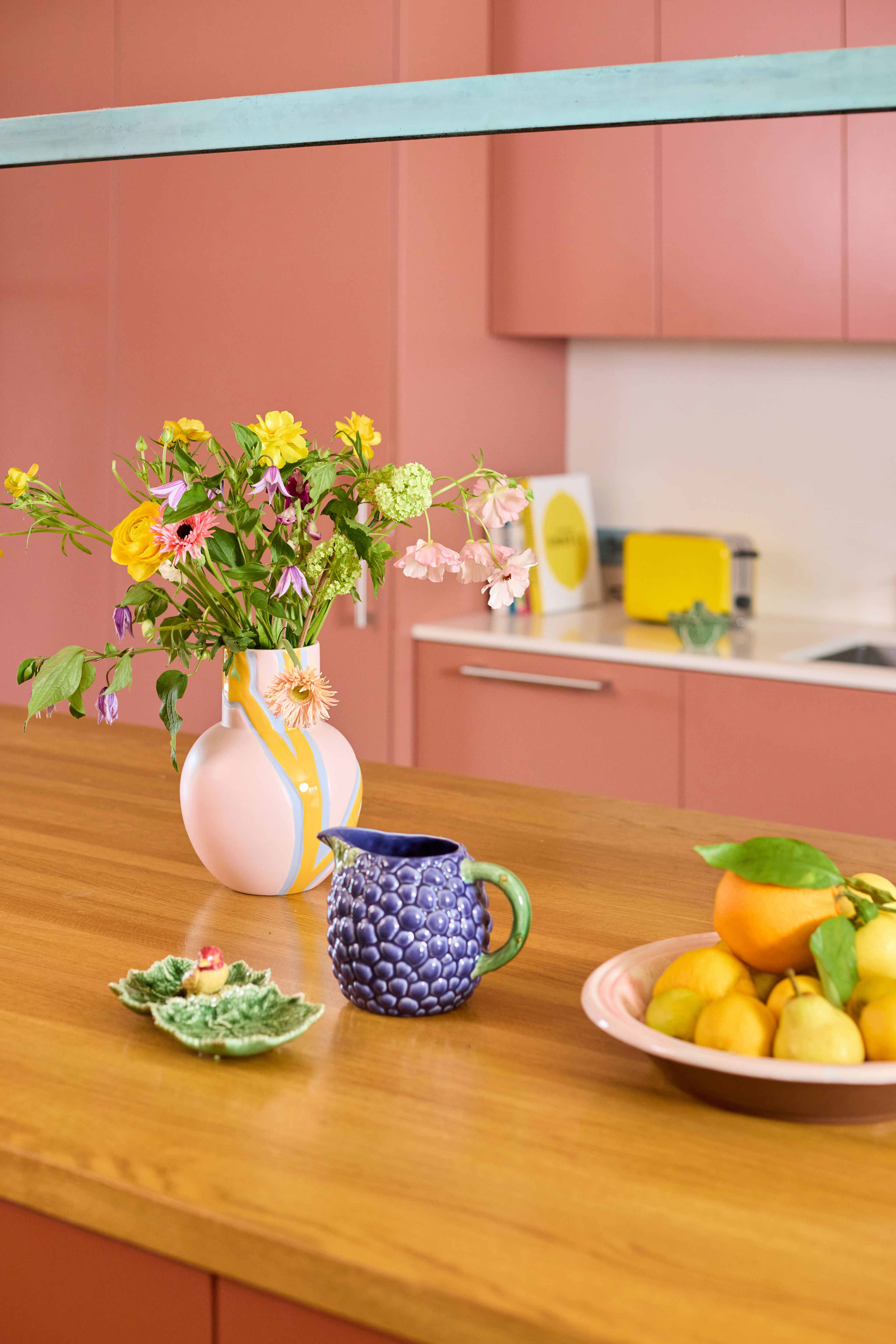Blomstervase, blå drueformet mugge og fat med sitrusfrukt på kjøkkenbenk i tre, terrakotta-kjøkken i bakgrunnen.