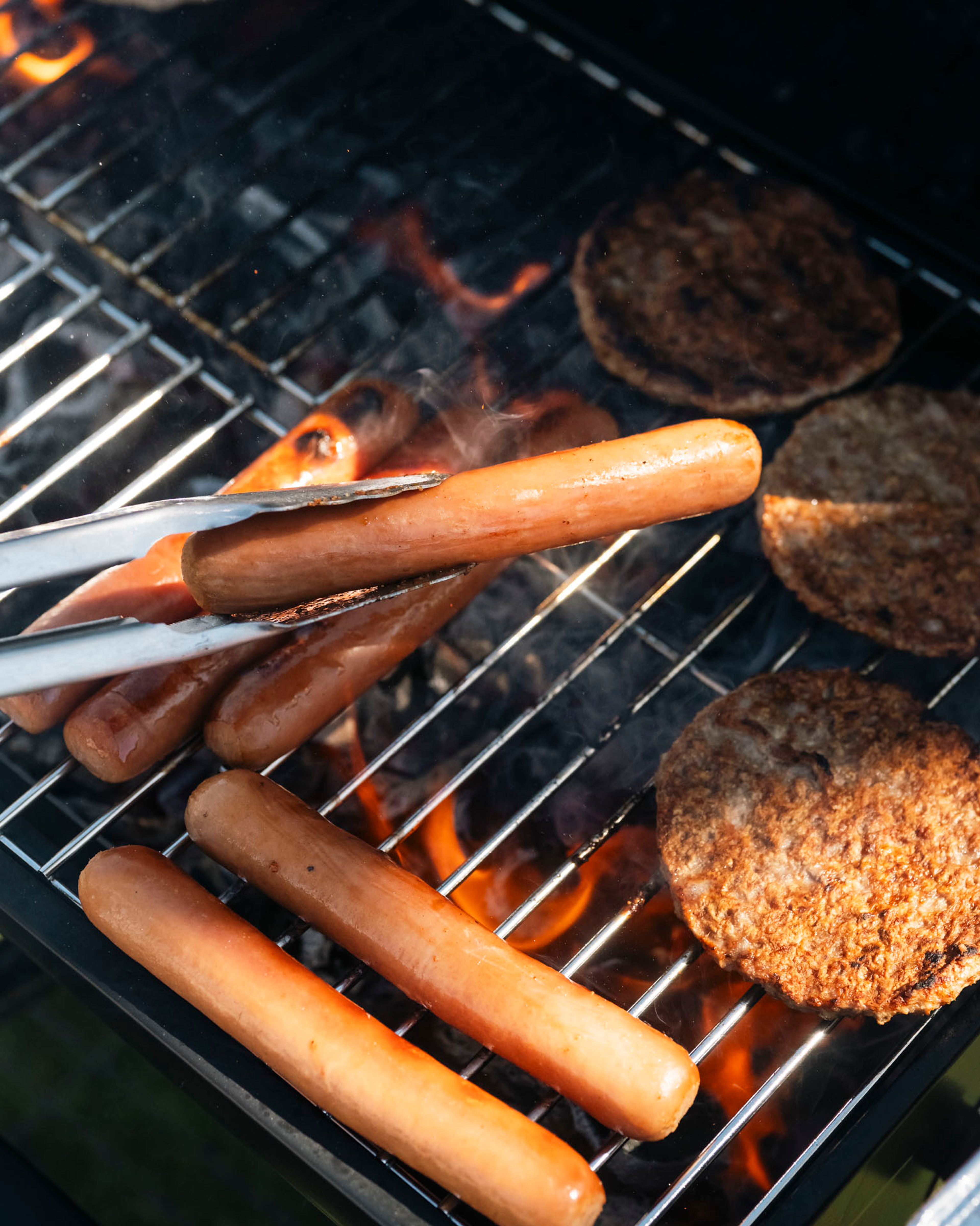 Pølser og hamburgere grilles over åpen flamme på en grill utendørs.