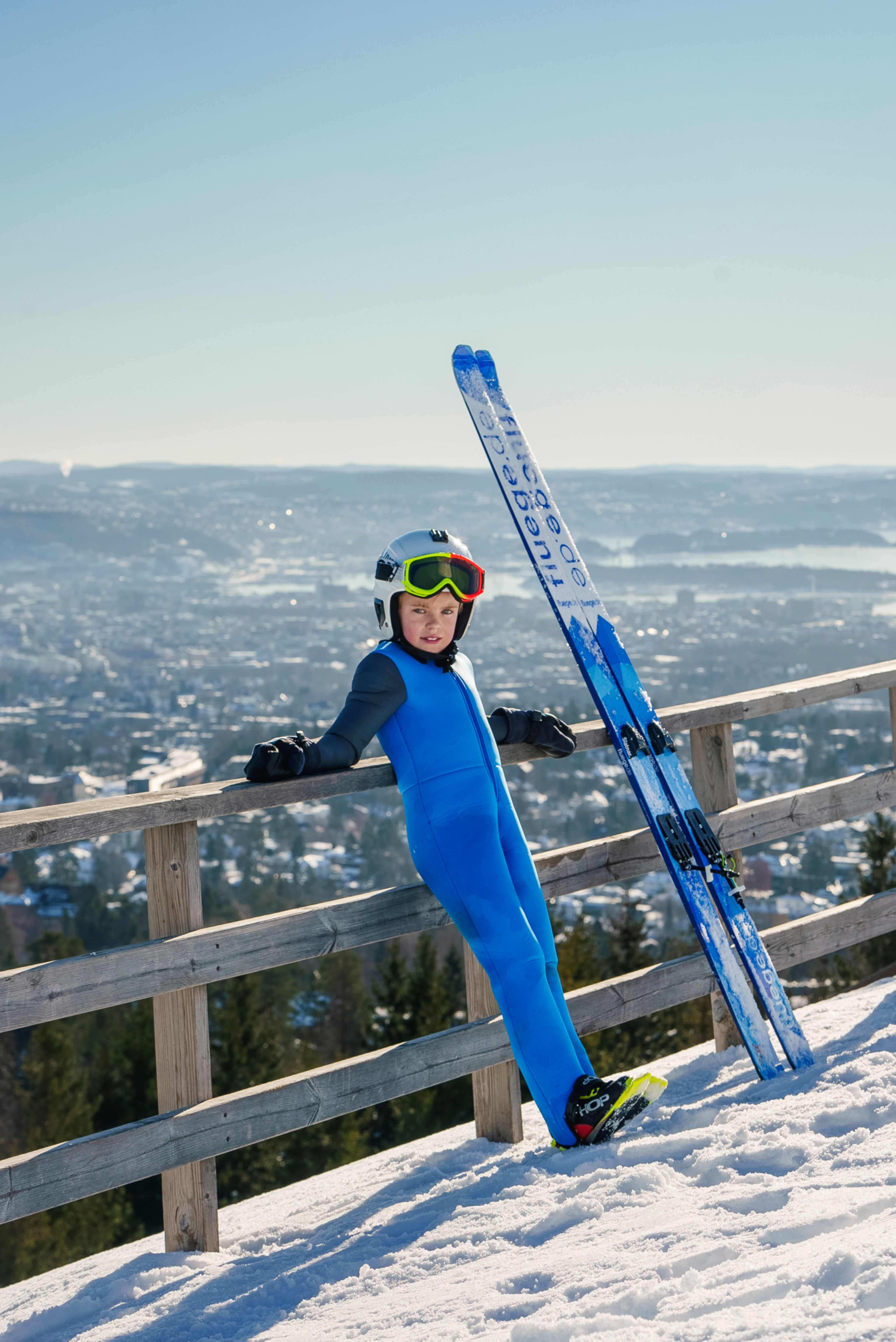 En unge i blå skidress lener seg mot et gjerde med utsikt utover Oslo-fjorden i bakgrunnen.