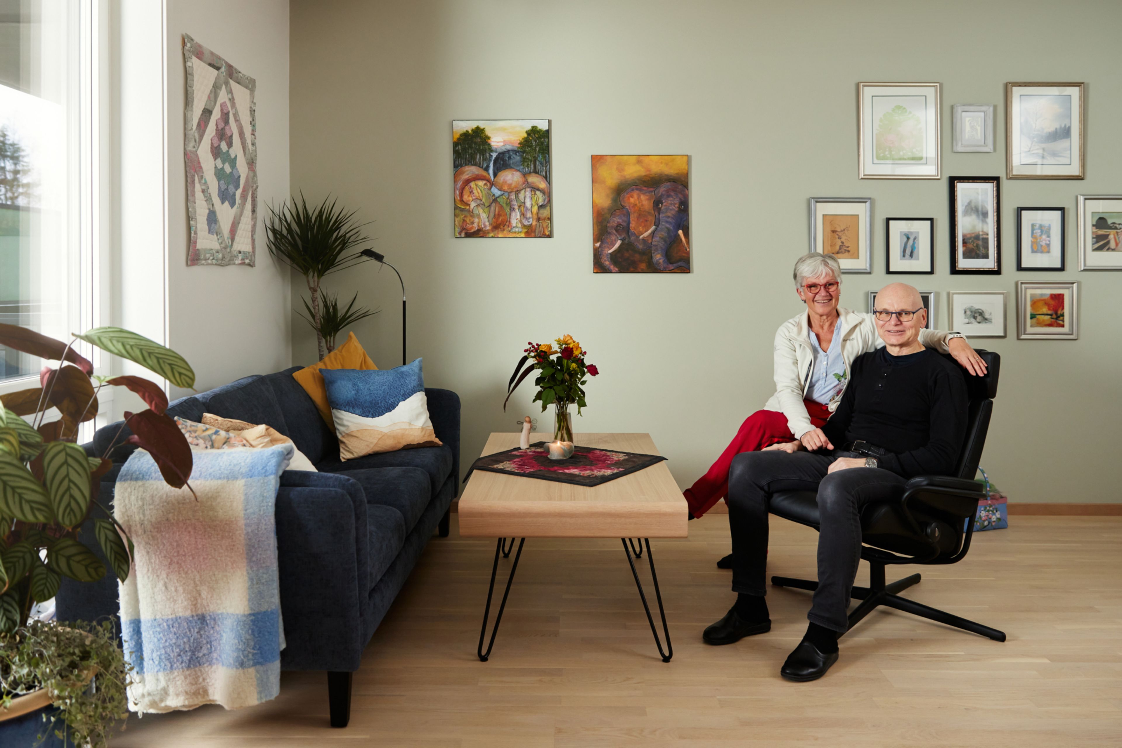 Foto av Solveig og Per som sitter i stua i leiligheten sin, med mange bilder på veggene.