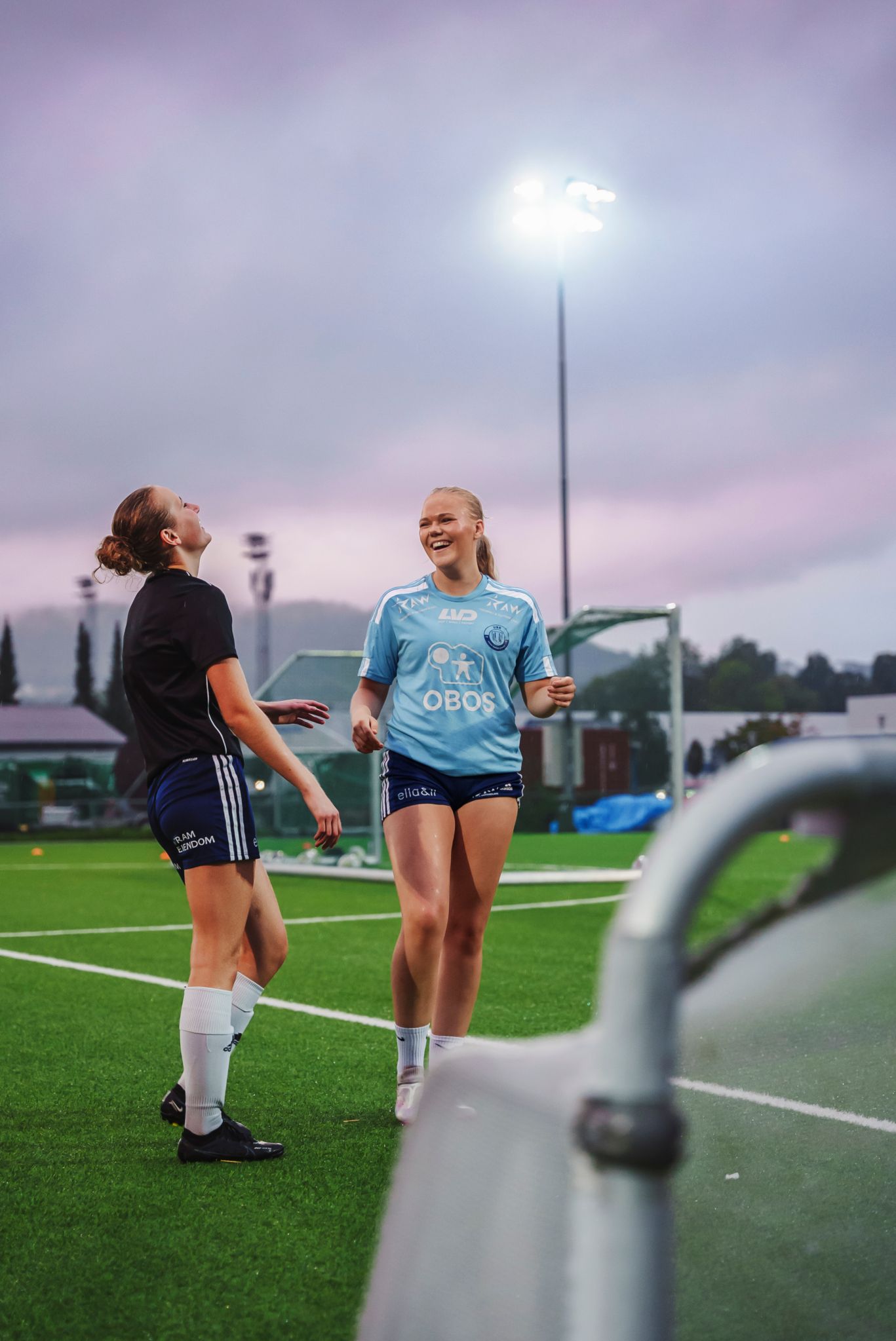 To spillere står og ler sammen på en kunstgressbane, med en lyskaster og en rosa himmel i bakgrunnen.