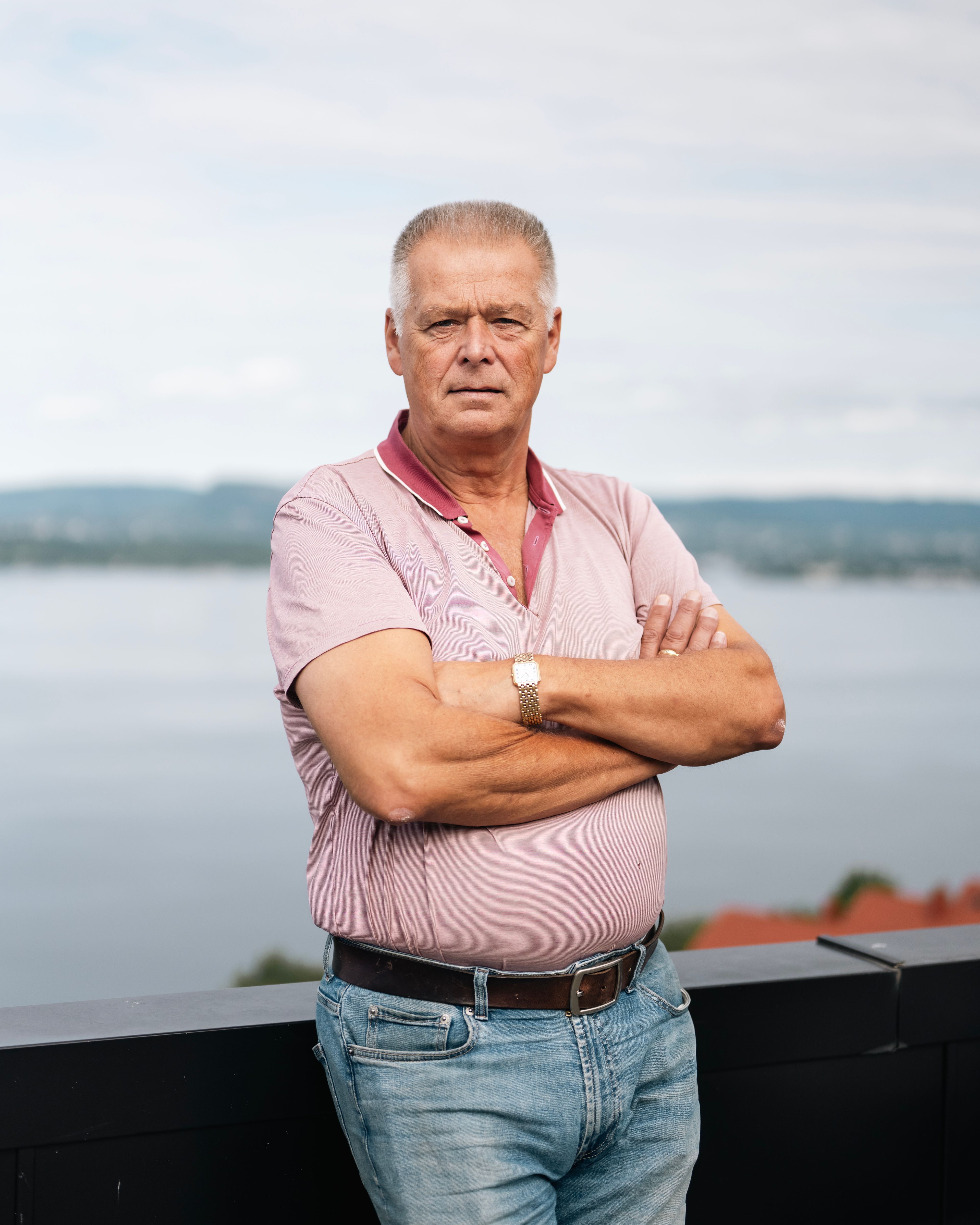 En mann står med armene i kors og lener seg inntil et rekkverk. Han har en lyserosa t-skjorte på seg. 
