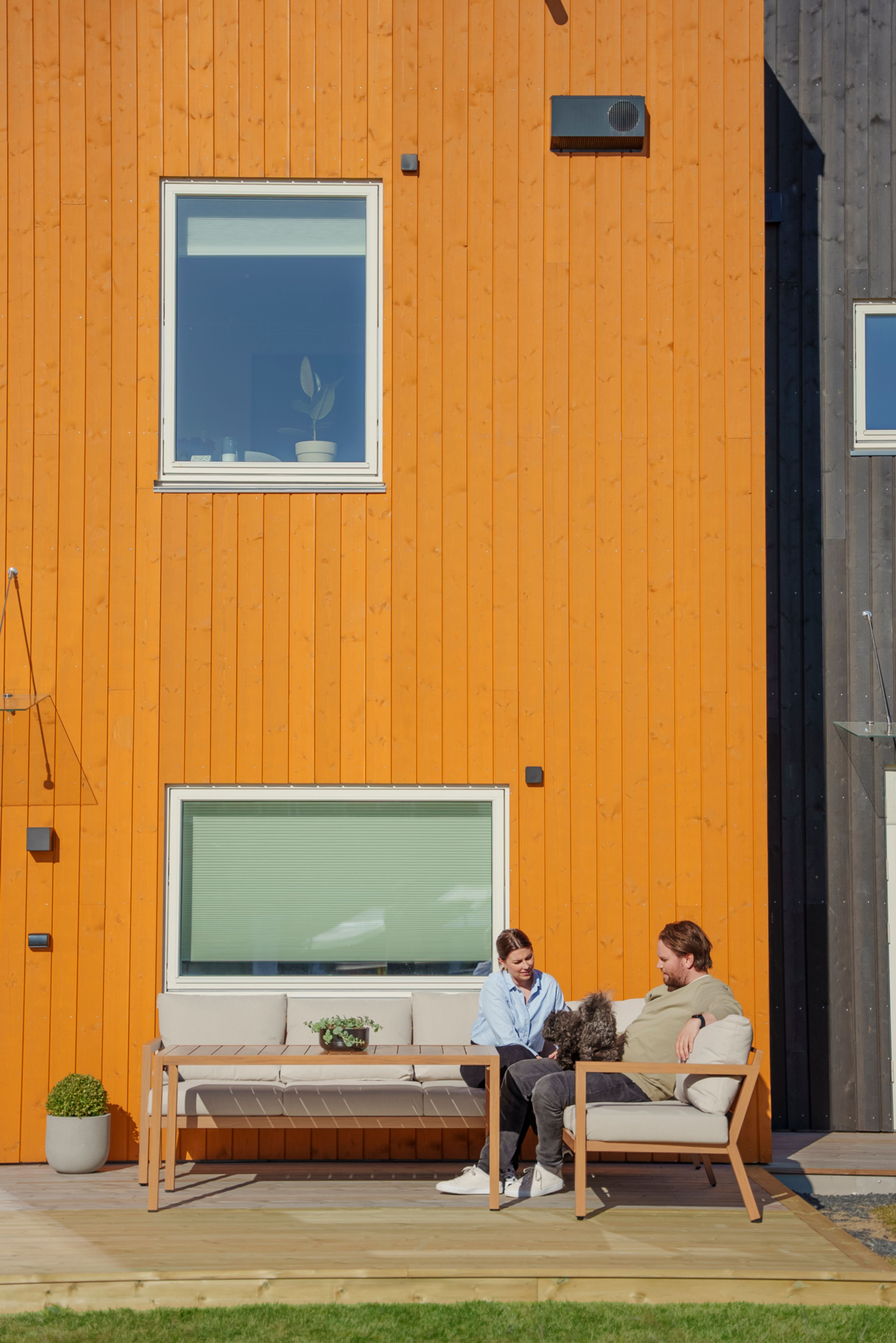 Stine og Martin sitter ute i en sofakrok mot den oransje ytterveggen på rekkehuset. Hunden Joa sitter mellom dem.