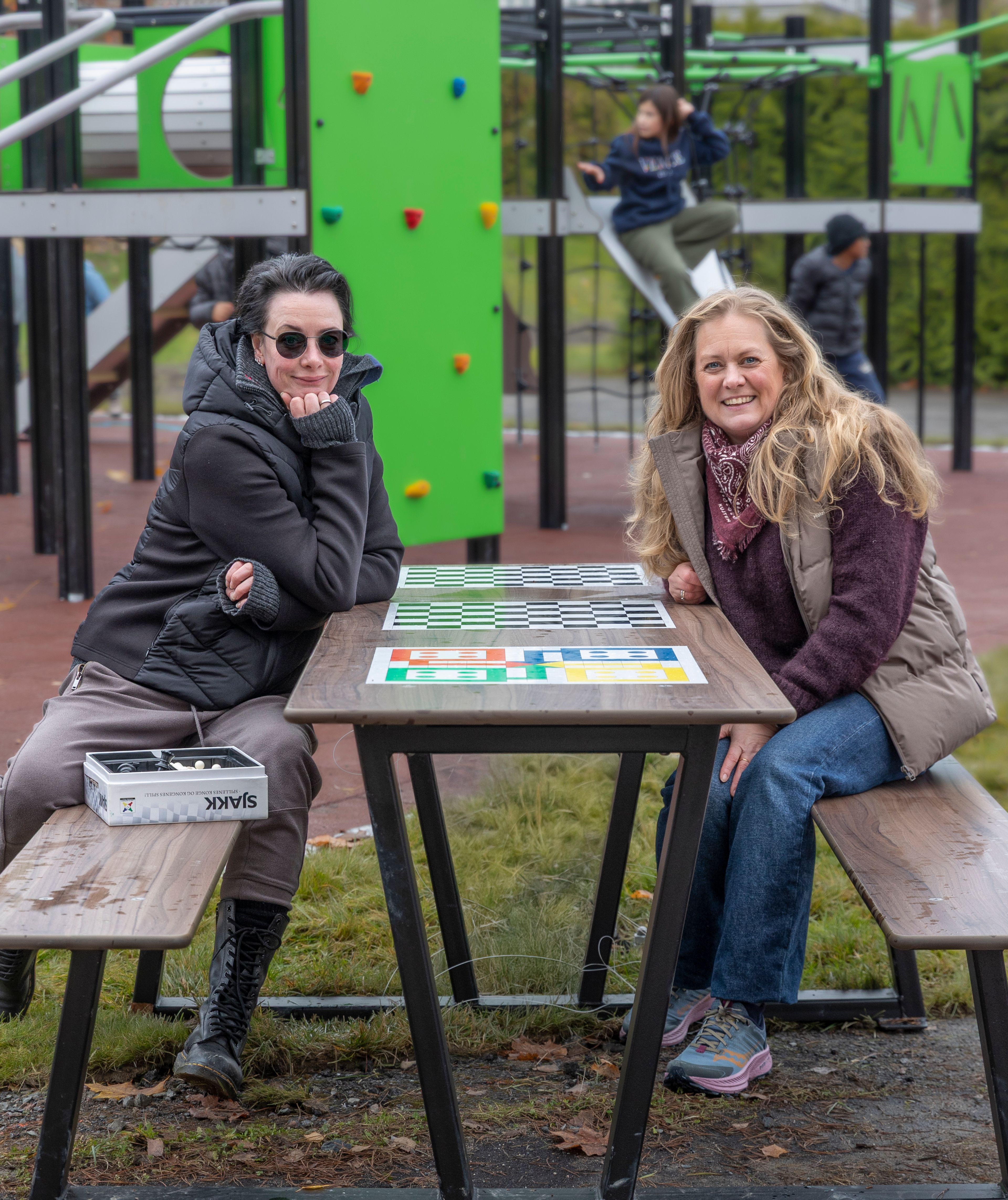 To kvinner sitter ved benk på en lekeplass