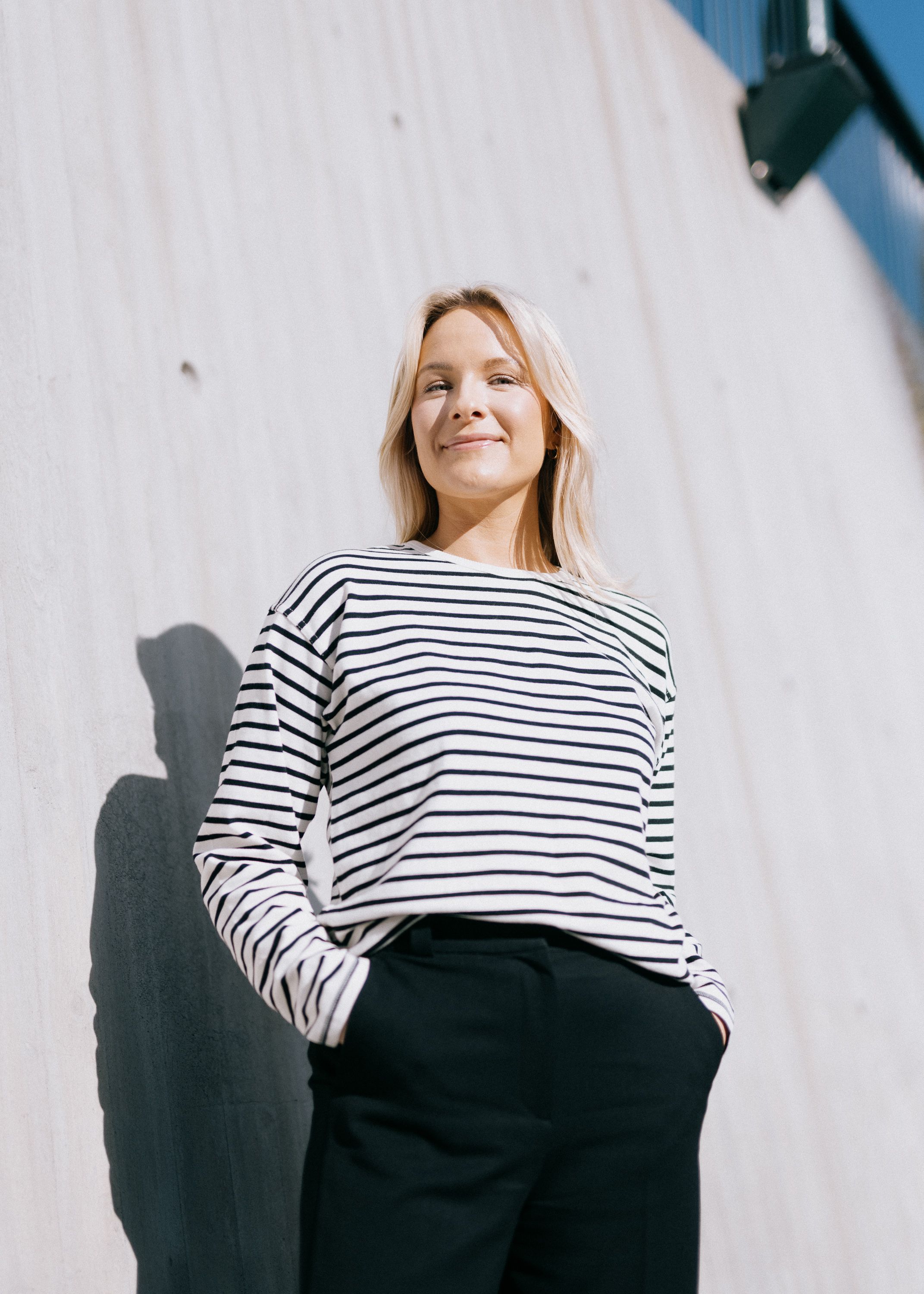 Foto av Brita som står framfor en murvegg. Hun har på seg en svart og hvit-stripete genser.