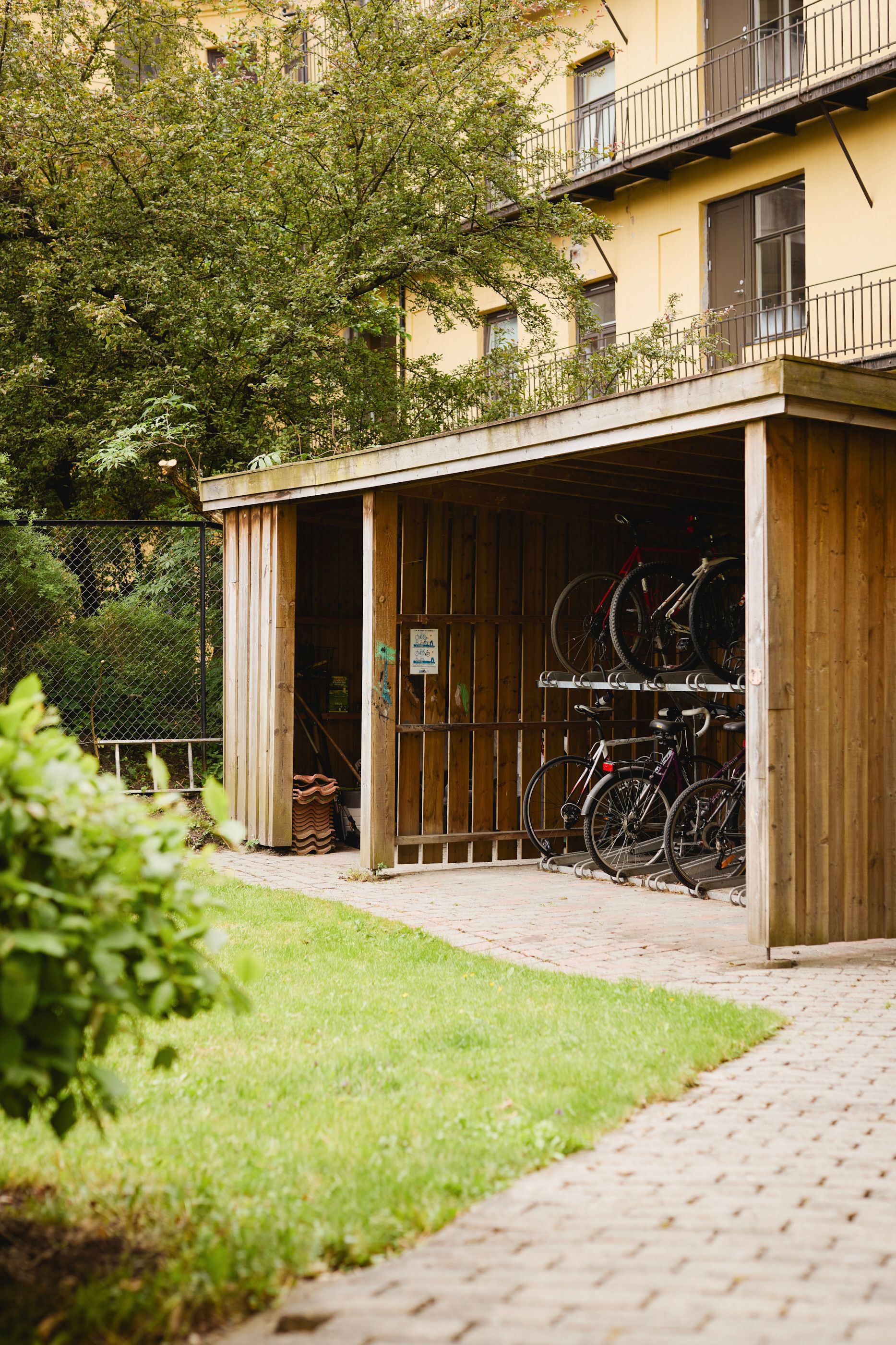 Utendørs sykkelskur i tre med flere sykler parkert i stativer, omgitt av grønt gress og trær foran en bygård.