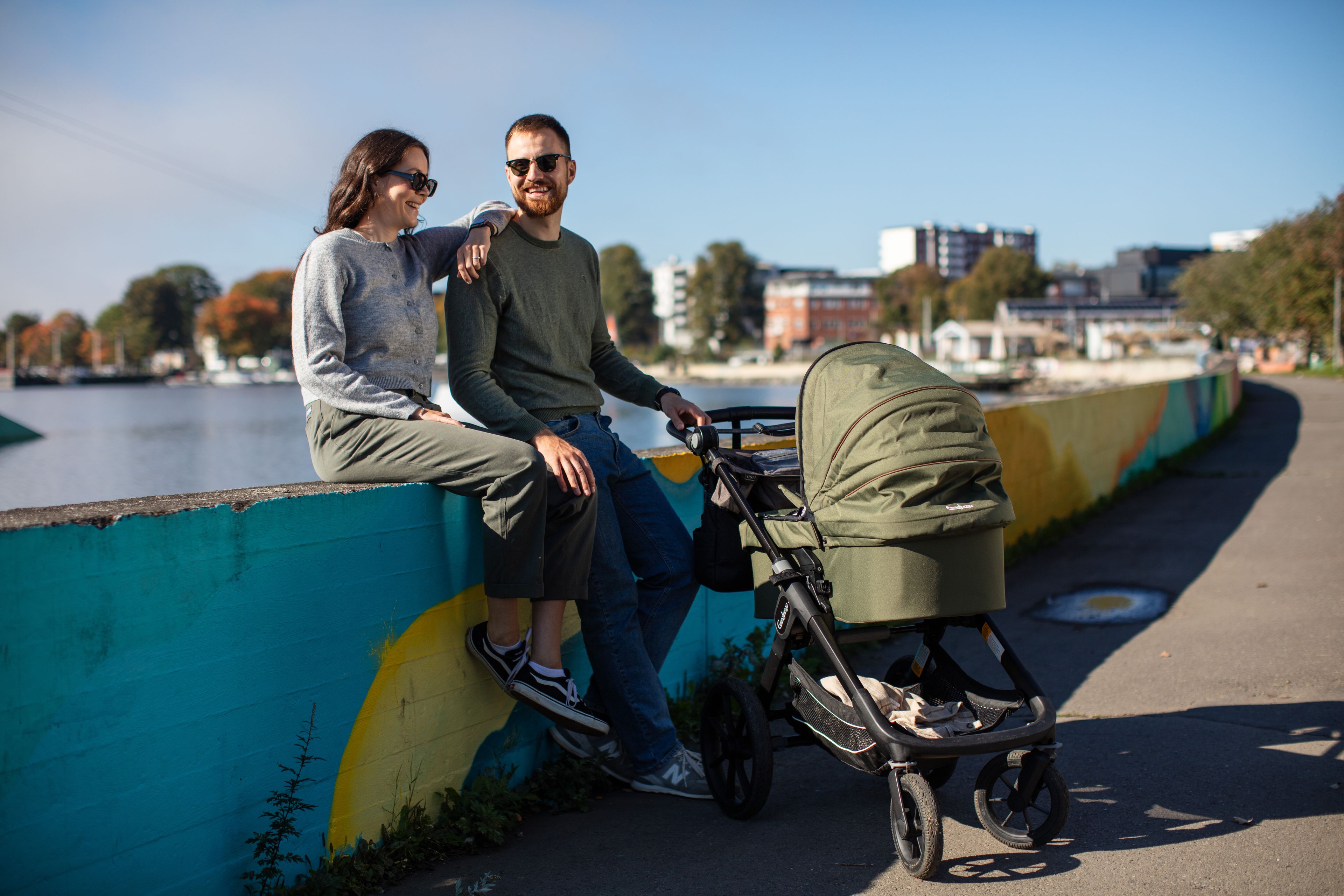 Et par som sitter på en lav vegg med barnevogn og vannet i Hamar i bakgrunnen