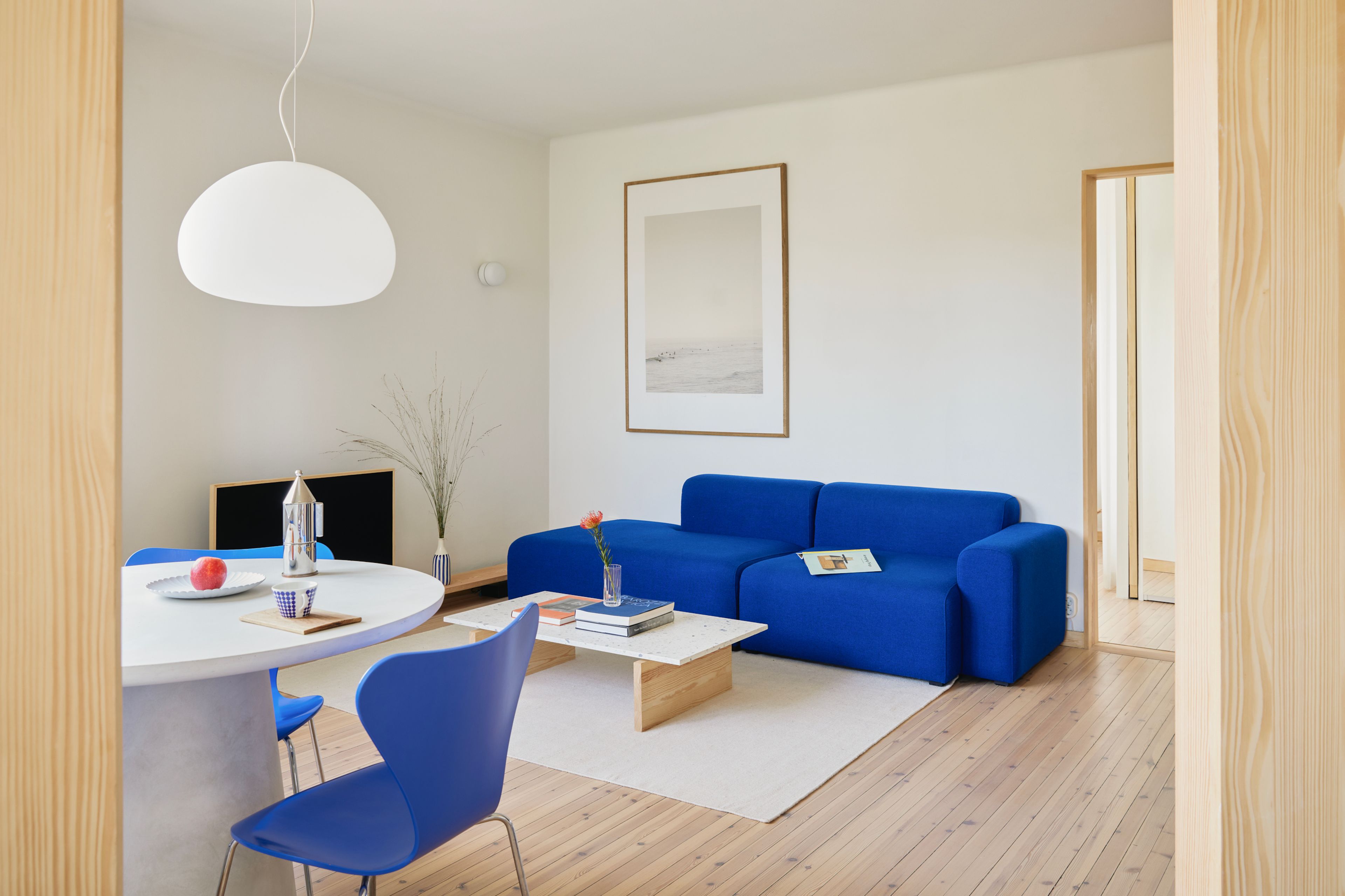 En blå sofa i en stue, den står inntil en hvit vegg. Foran sofaen er det et lite bord. Vi ser også et spisebor med en blå spisestol.