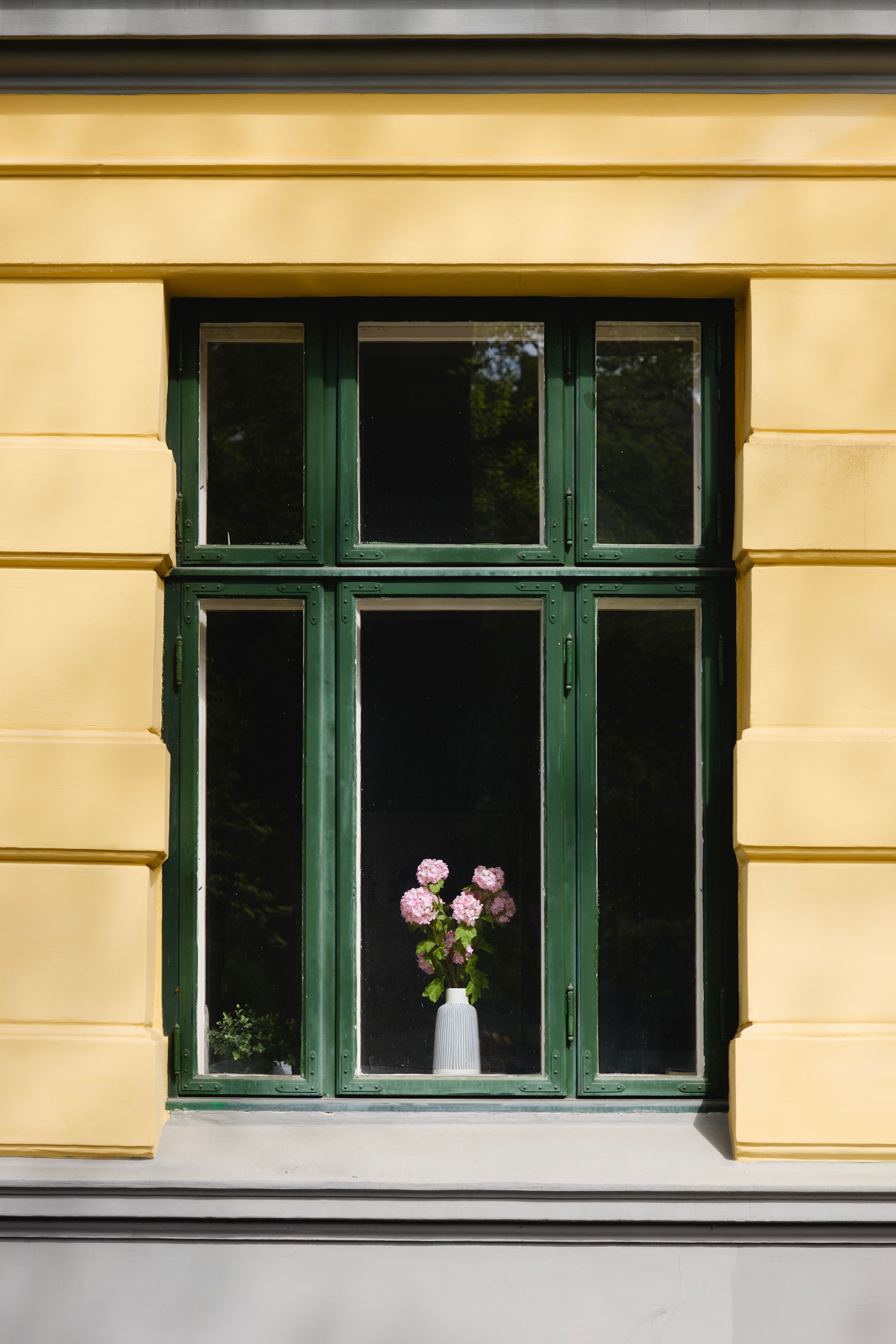 Vindu tilhørende en gammel bygård. Vinduskarmene er grønn og det står en vase med rosa blomster i vinduskarmen.