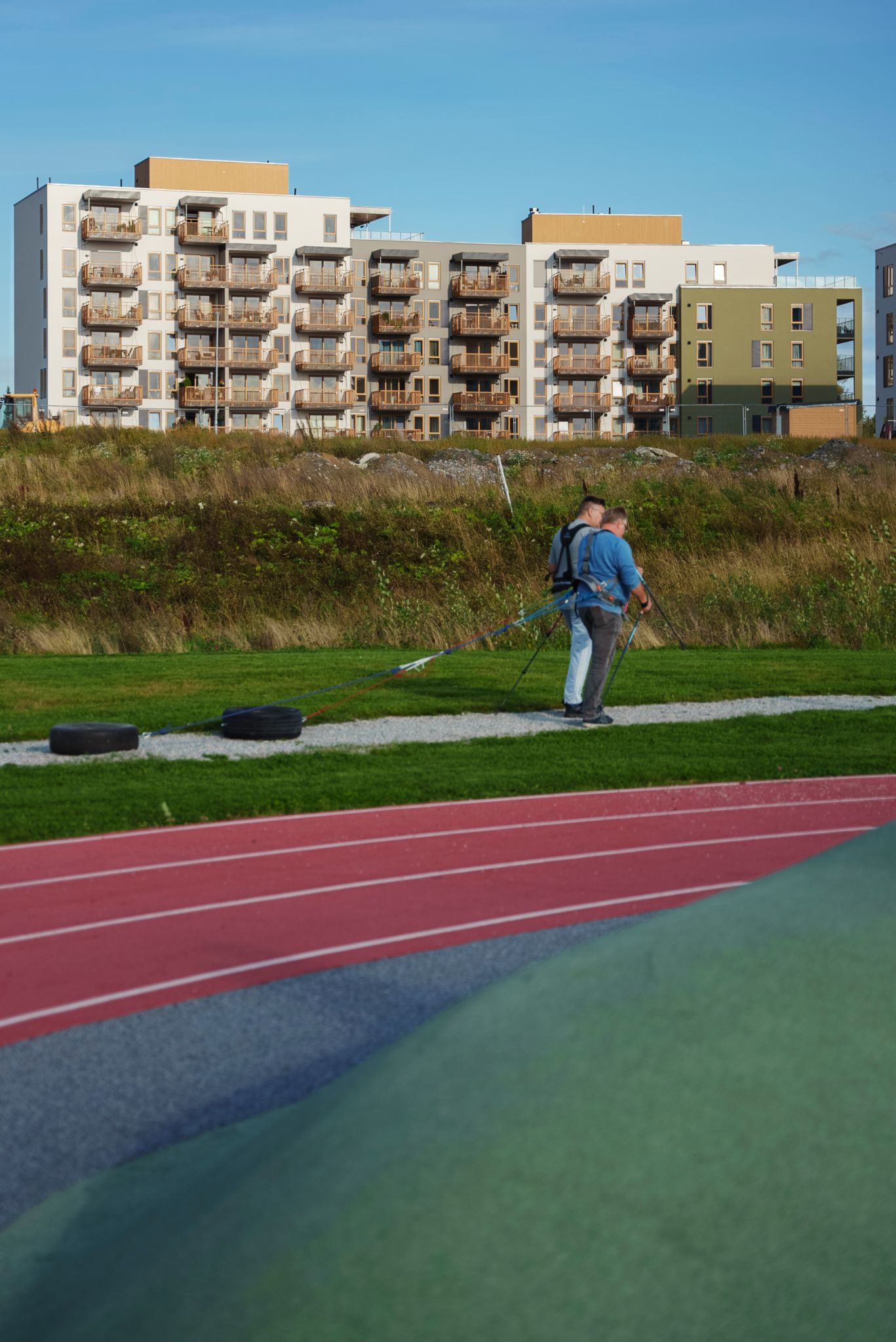 Bilde av en løpebane med Asper Hageby i bakgrunnen, hvor to personer går tur med staver og drar et dekk bak seg.