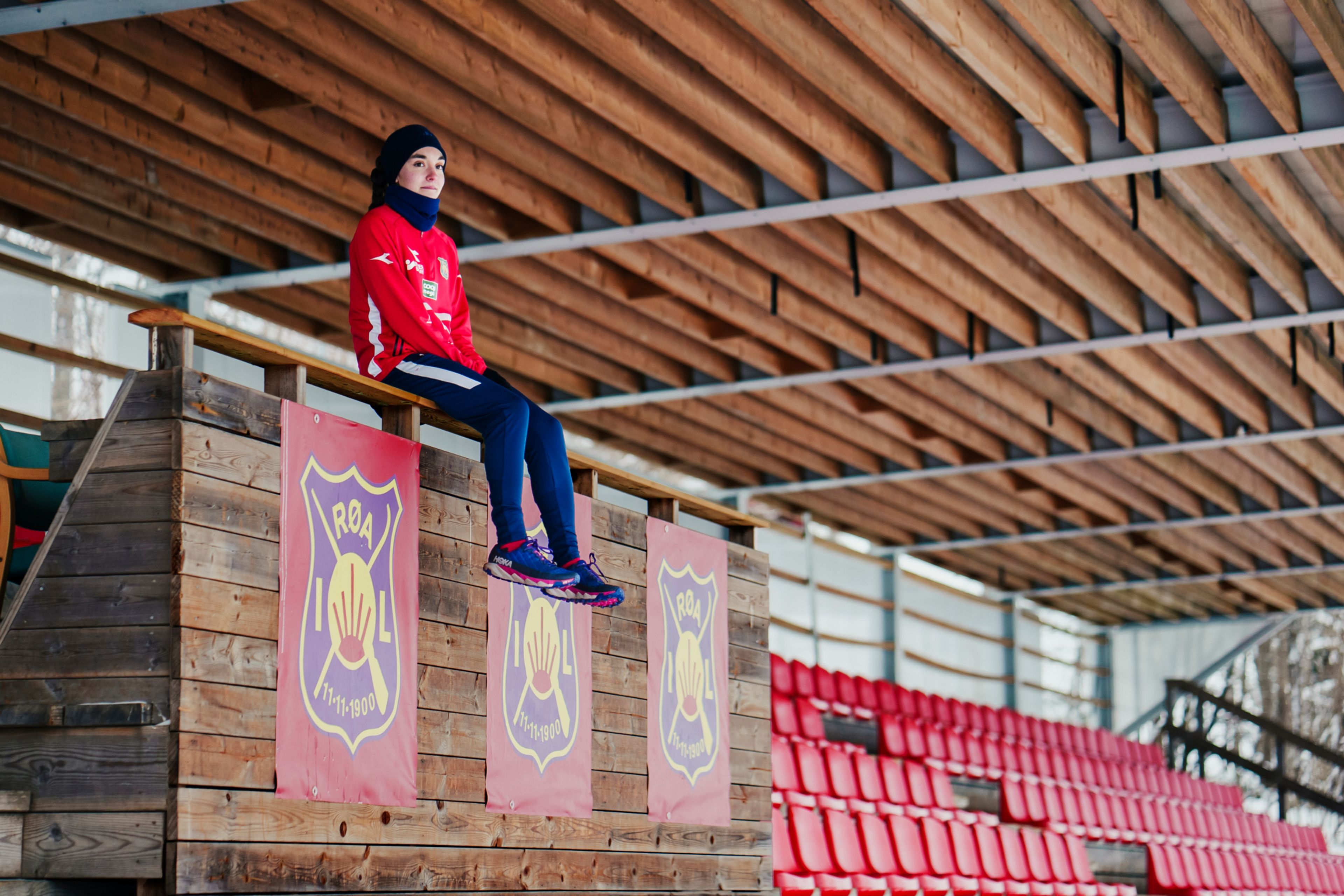 Sara sitter på tribunen og ser utover fotballbanen. Hun har på seg rød genser, blå bukse og svart pannebånd.