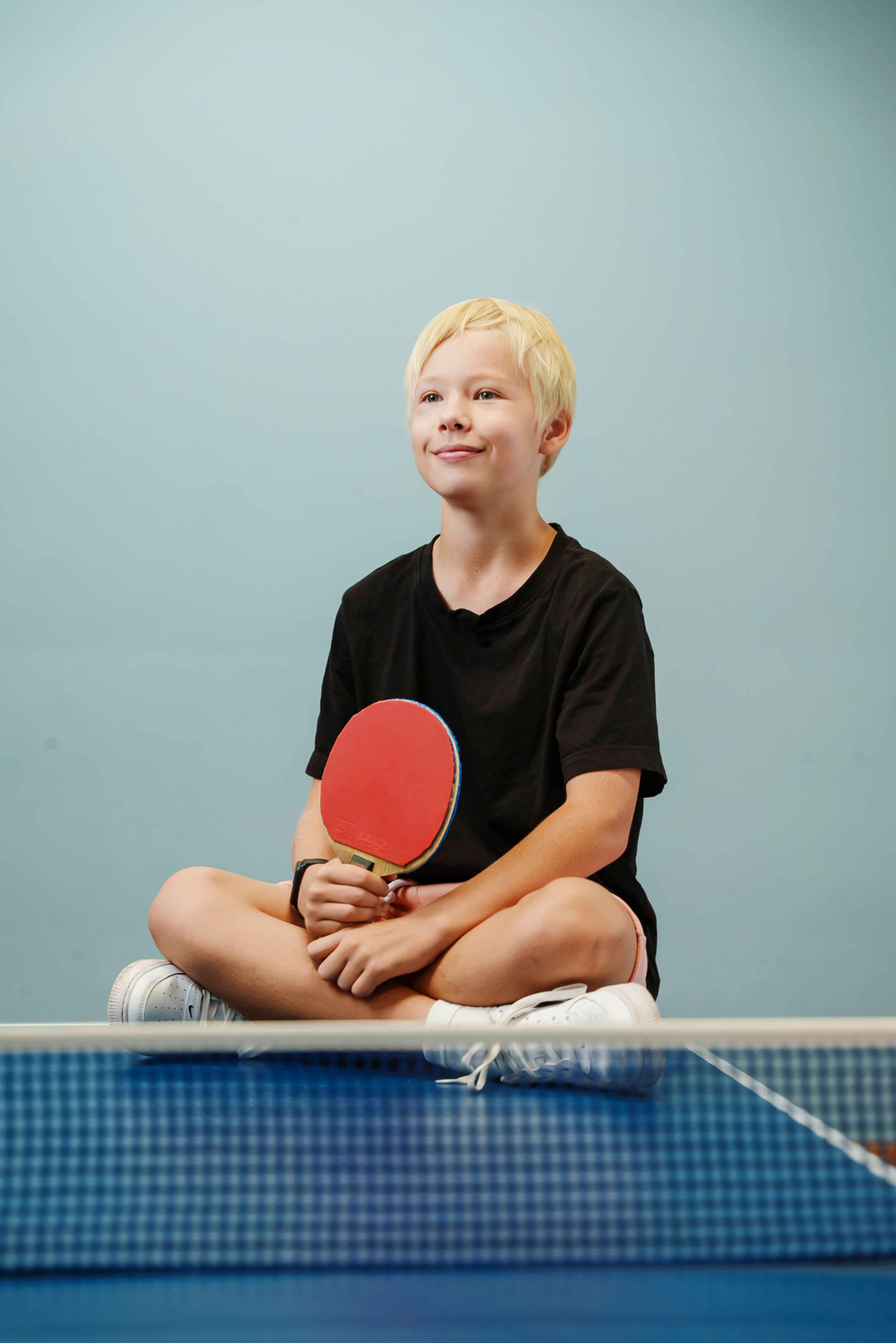 En yngre gutt sitter på et bordtennisbord og holder i en rackert.
