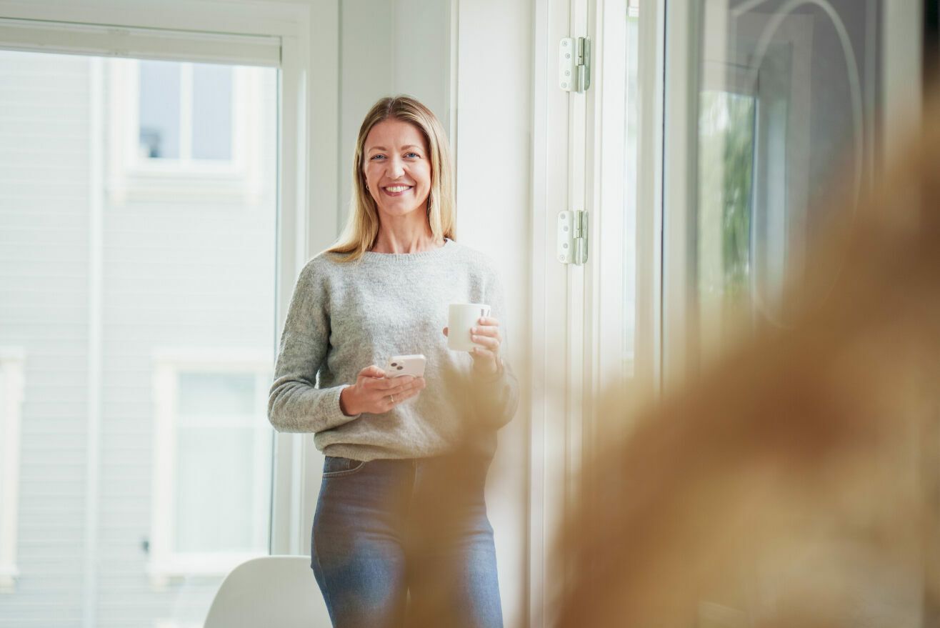 Forbrukerfrue smiler med en kaffekopp og mobilen i hånda.