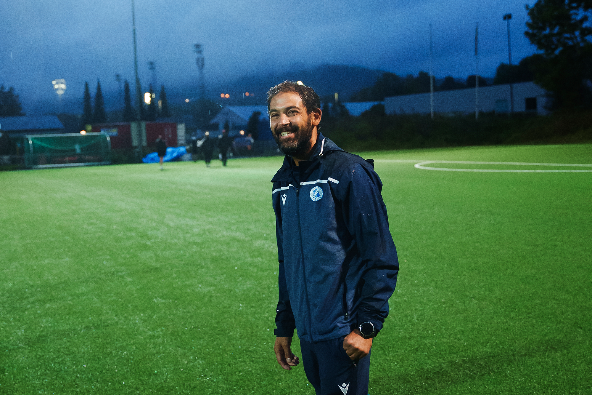 Hovedtrener Fahed står og smiler mot kamera på fotballbanen i kveldsmørket.