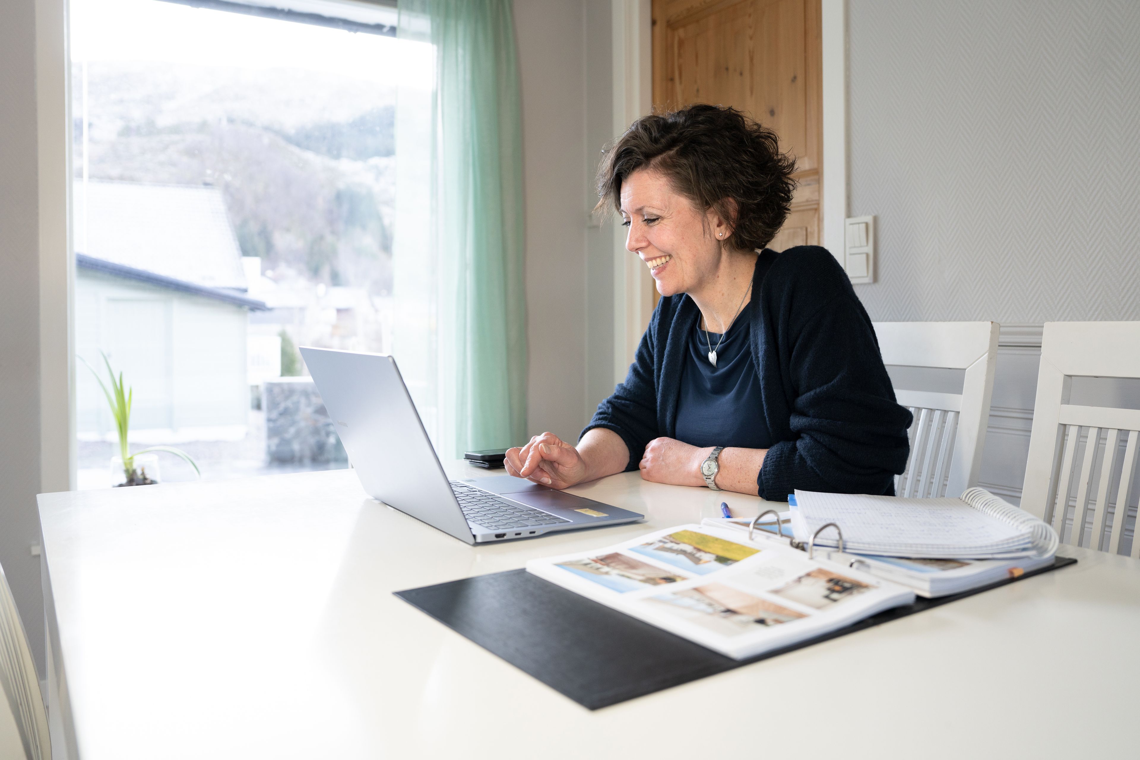 Marion Vik sitter ved PC-en sin og smiler. Ved siden har hun tegninger av huset i en mappe.