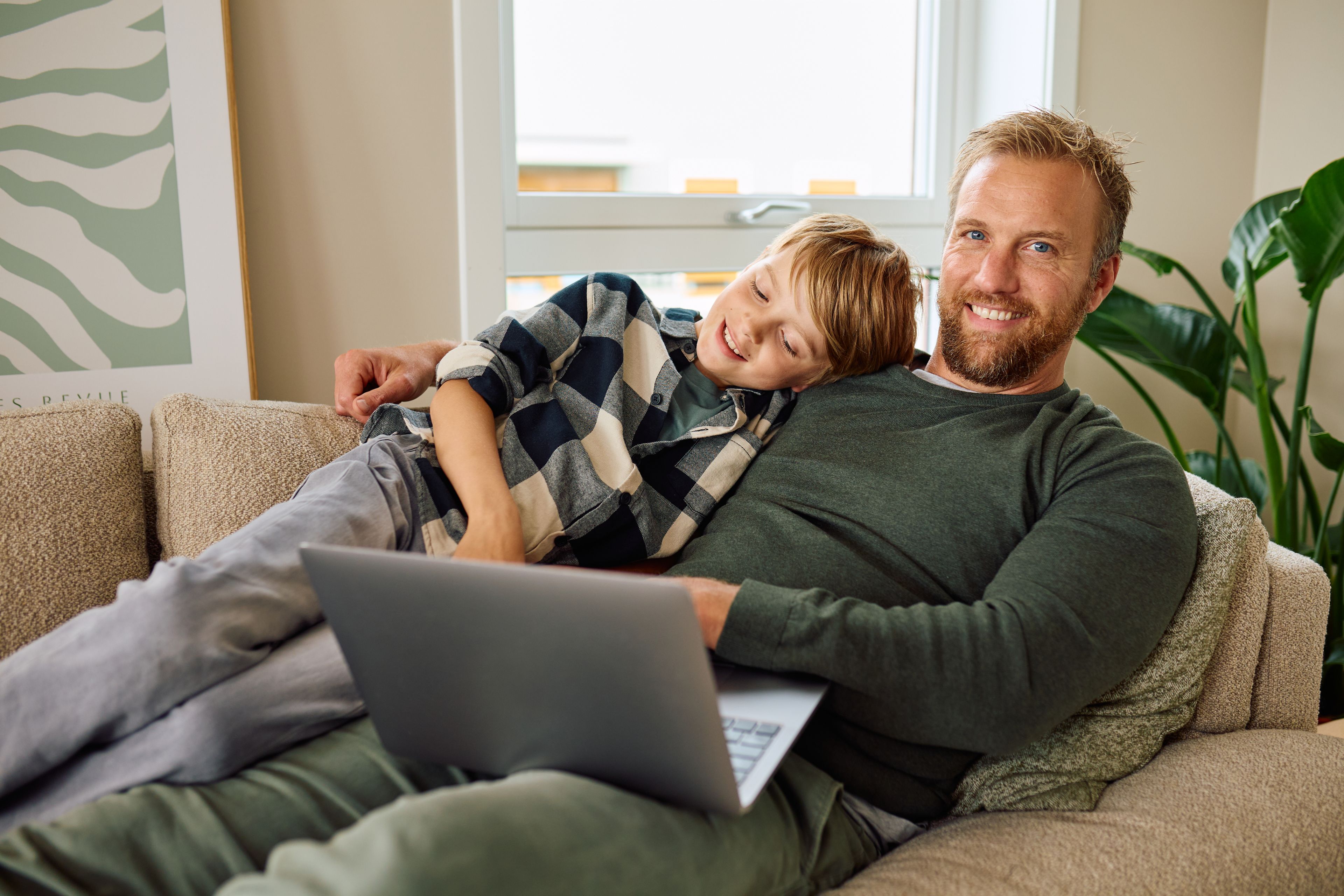 En mann ligger med  PC-en i fanget, på sofaen. Han smiler. En gutt ligger i armkroken hans.