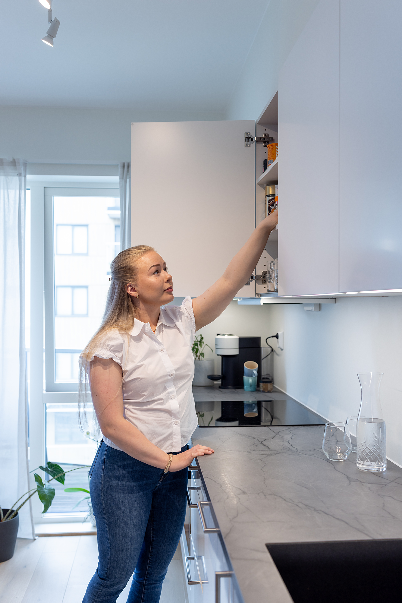Amanda står på kjøkkenet og lener seg opp i en av hyllene over kjøkkenbenken. Kjøkkenet er i lyse farger.