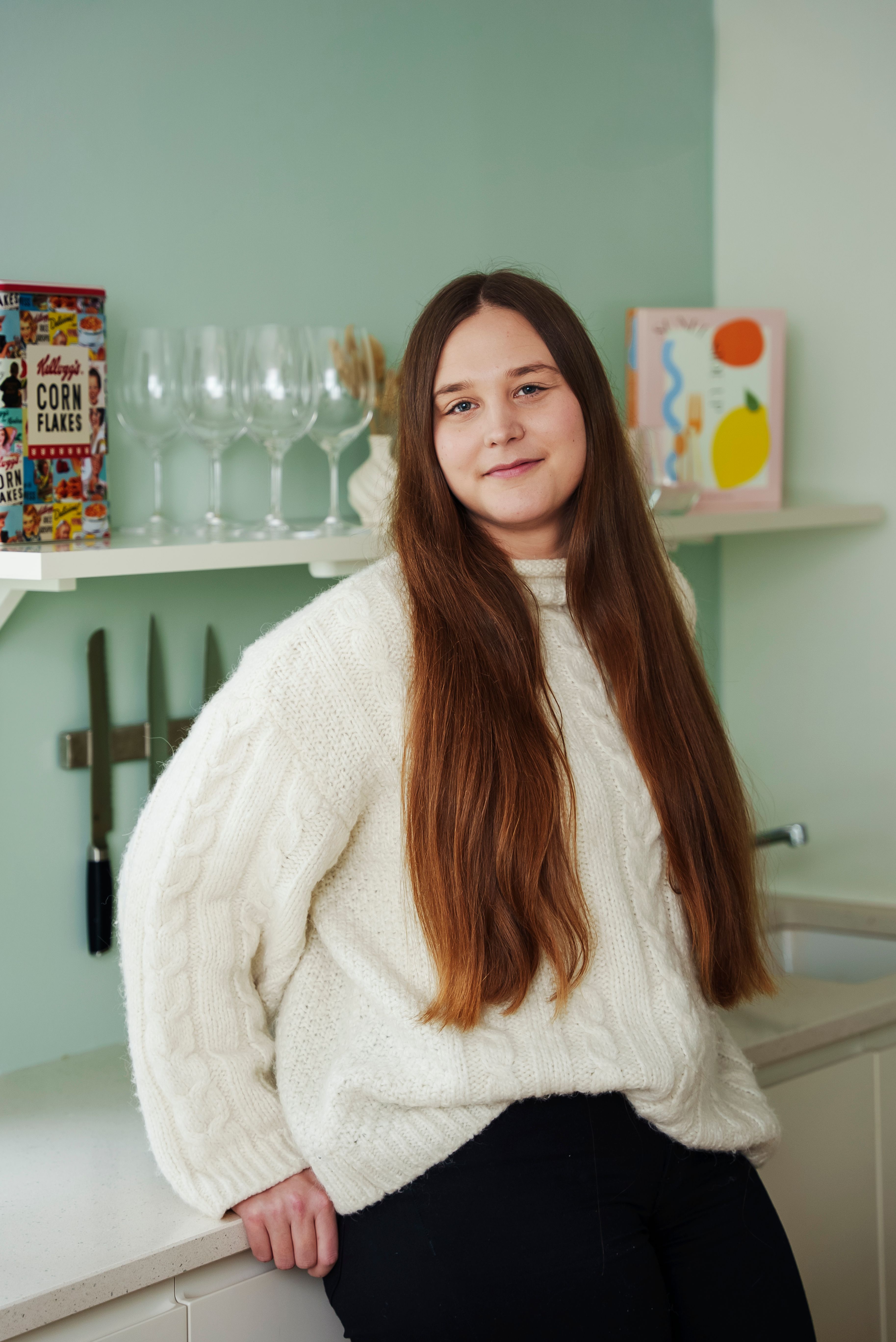 Kristine Istad smiler forsiktig mot kamera mens hun lener seg mot kjøkkenbenken på et lyst, moderne kjøkken