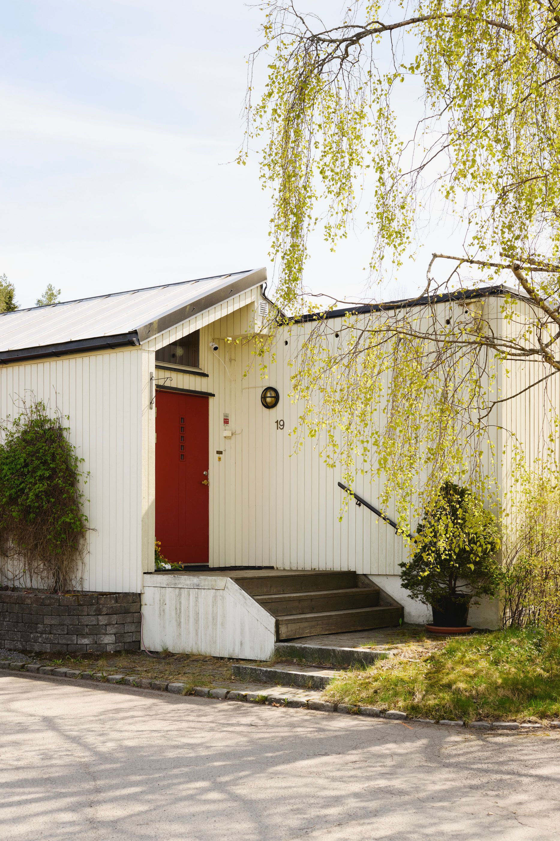 Inngangsparti til en hvitmalt bolig med rød dør, omgitt av grønne vekster og en overhengende bjørk.