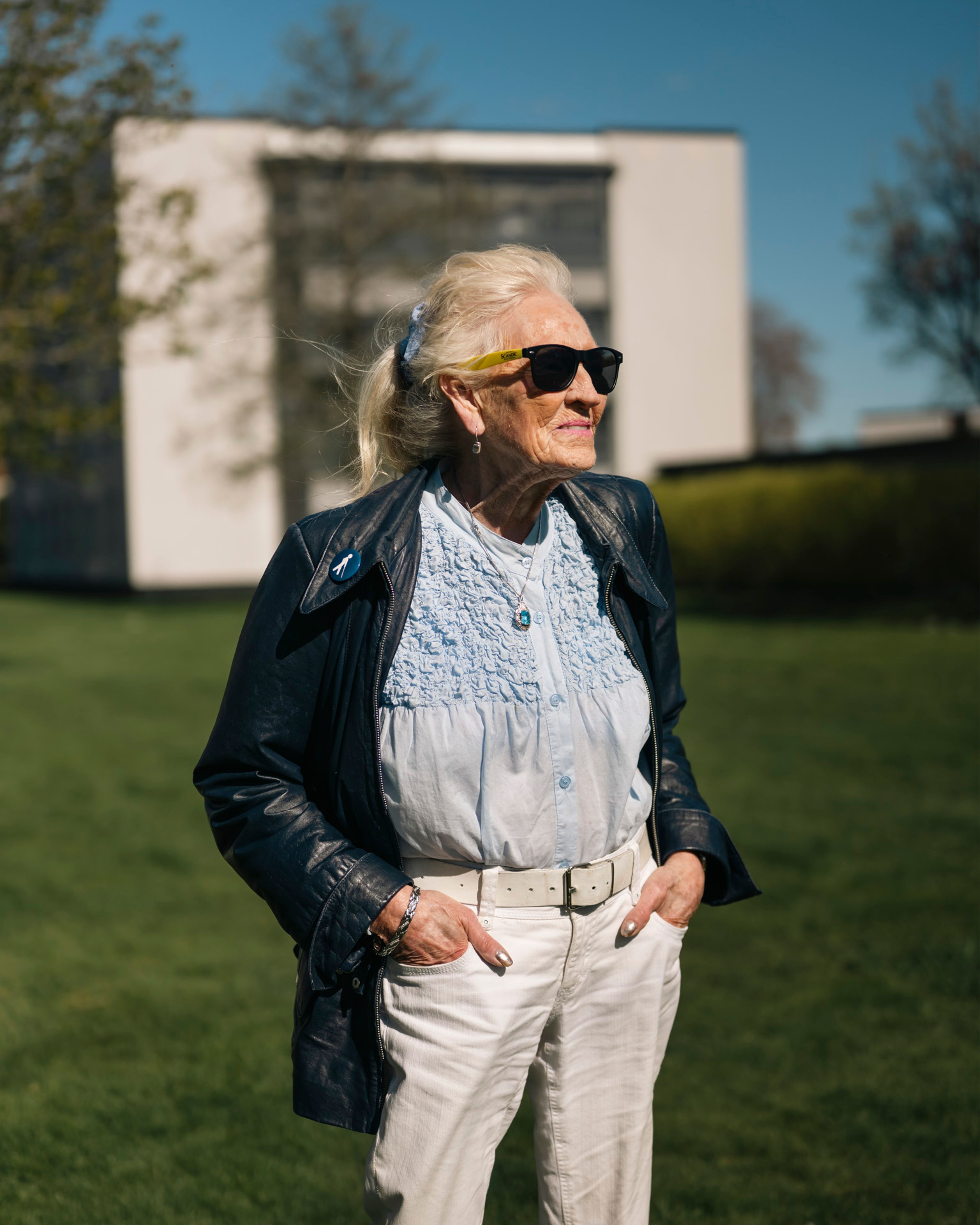 Eldre kvinne med langt hvitt hår, solbriller og mørk skinnjakke står på gressplenen foran de hvite blokkene.