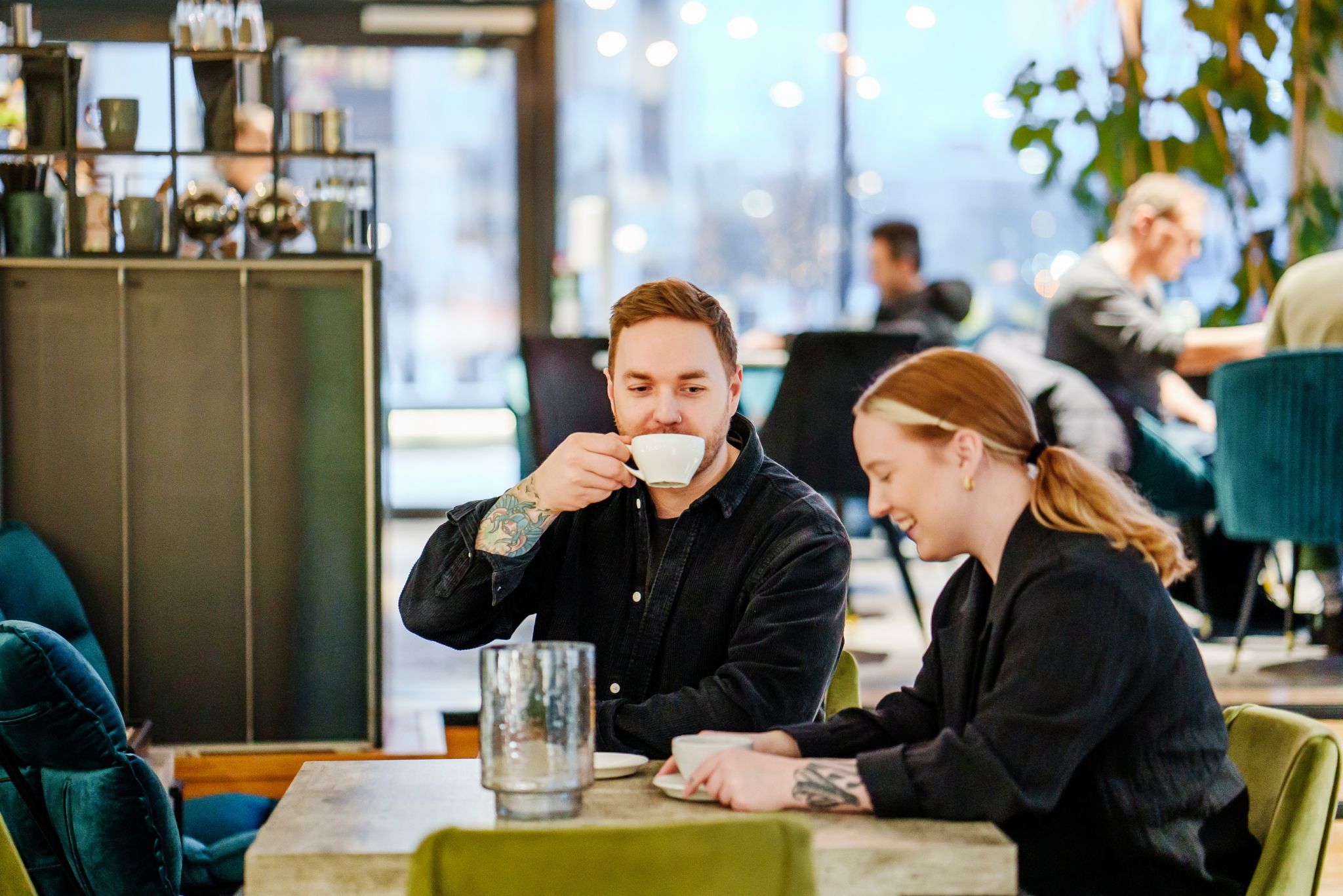 En mann og en kvinne sitter ved et bord på en kafé, han drikker av en kaffekopp.