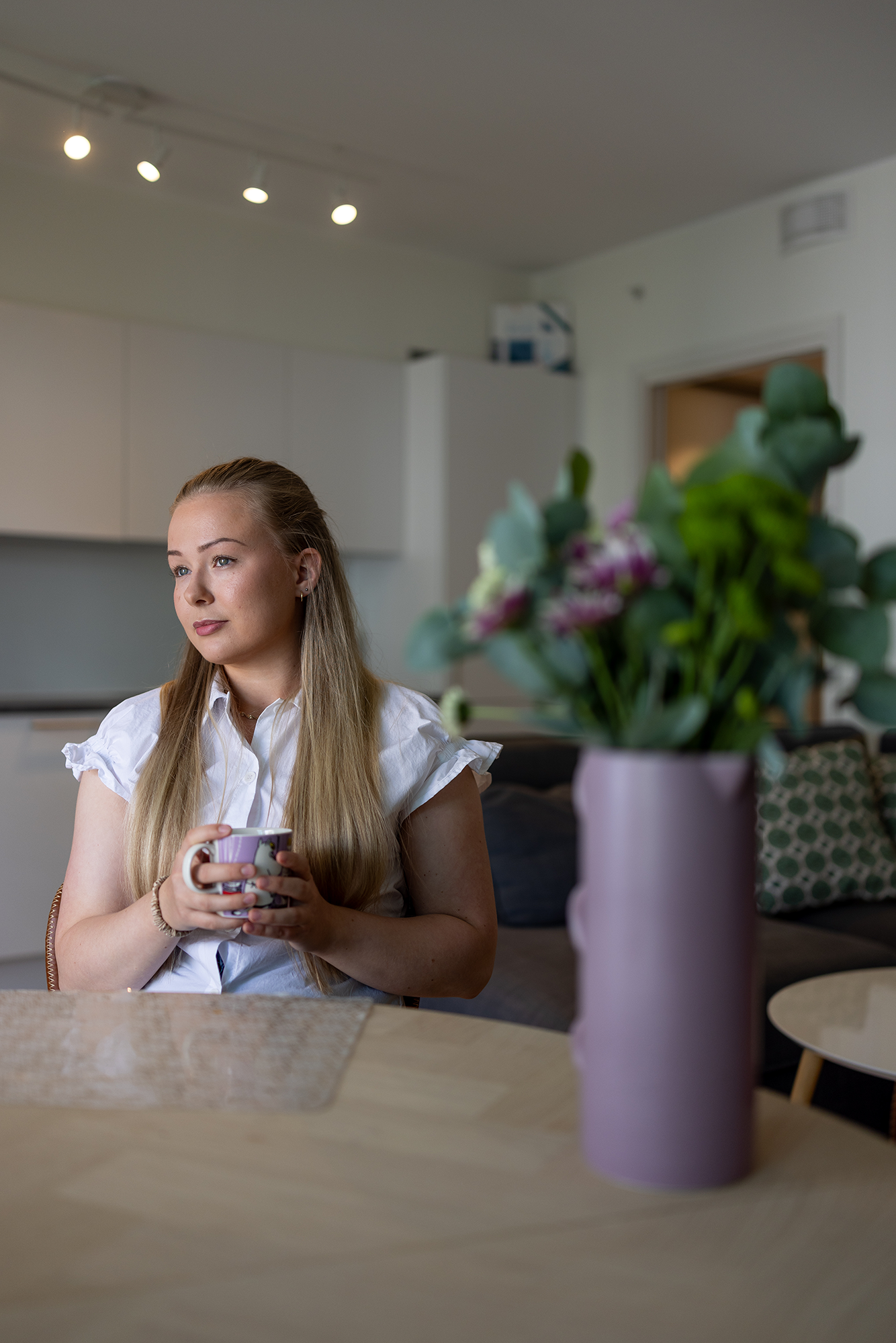 Amanda sitter ved stuebordet og ser ut vinduet, med en kaffekopp som hun holder med begge hender.