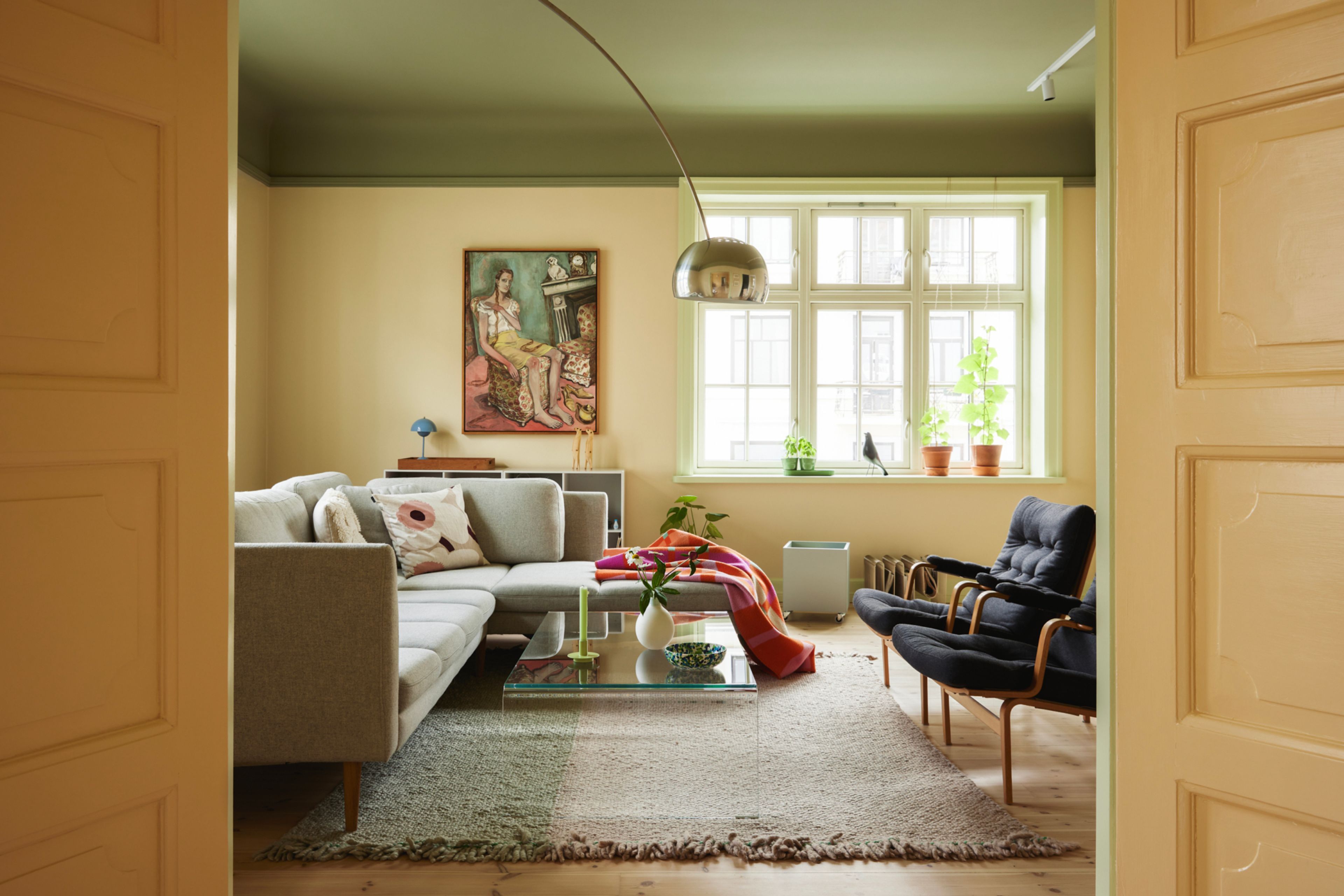 Stuen i leiligheten har en dus gulfarge på veggen og et stort vindu med sprosser. Sofaen står midt i rommet med to stoler midt imot.