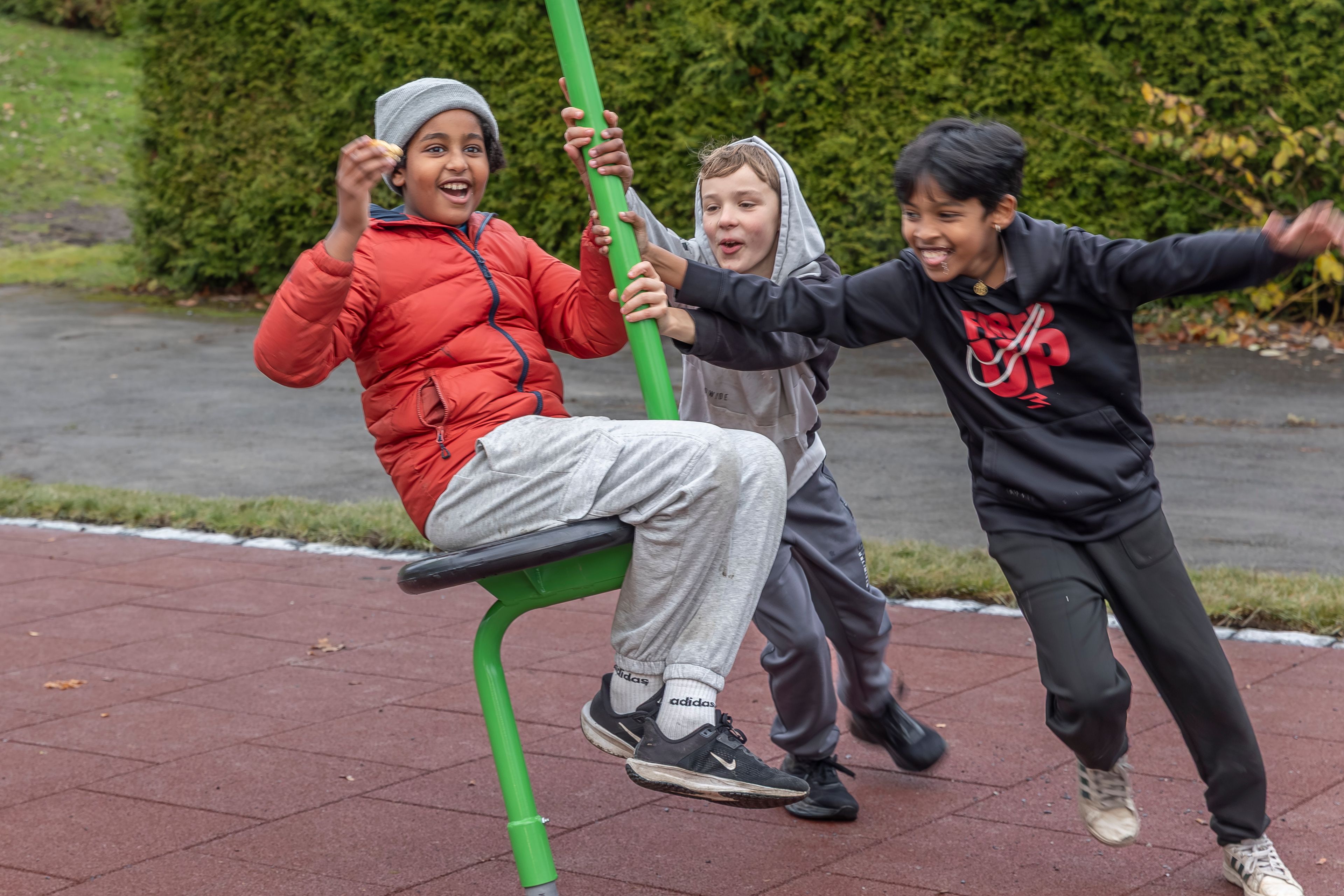 Tre barn leker på en grønn karusell på lekeplassen i Stovner borettslag, mens de ler og dytter hverandre for fart.