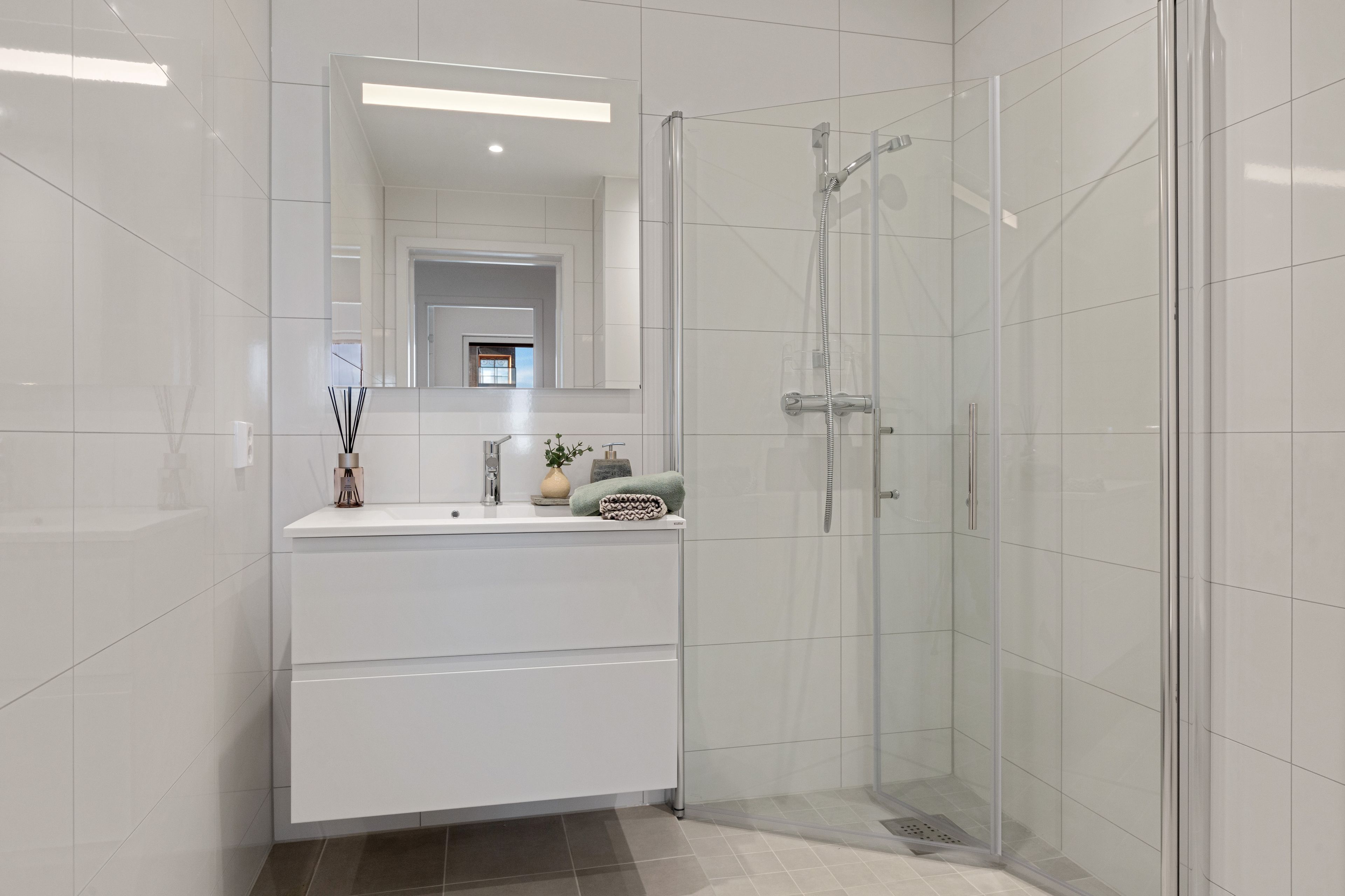 Bad i en leilighet i Furukrona innredet med servantskap med vask og dusjhjørne med dusjvegger i glass som kan foldes inn mot veggen. Badet har hvite fliser på veggen og beige, mindre fliser på gulvet.