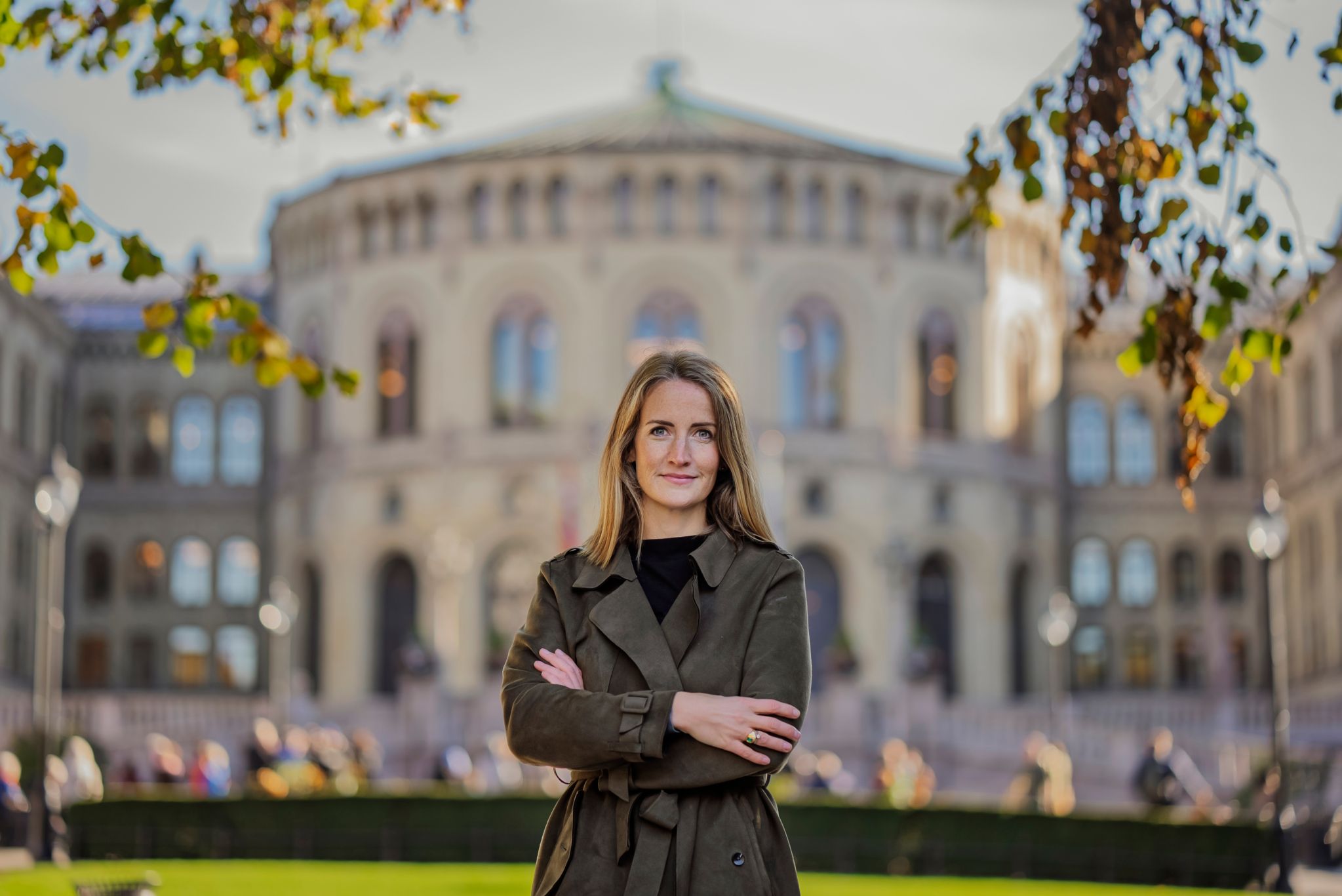 Ingjerd står i en gråbrun kåpe med armene i kors foran Stortinget. Det er sol og grønt ute.
