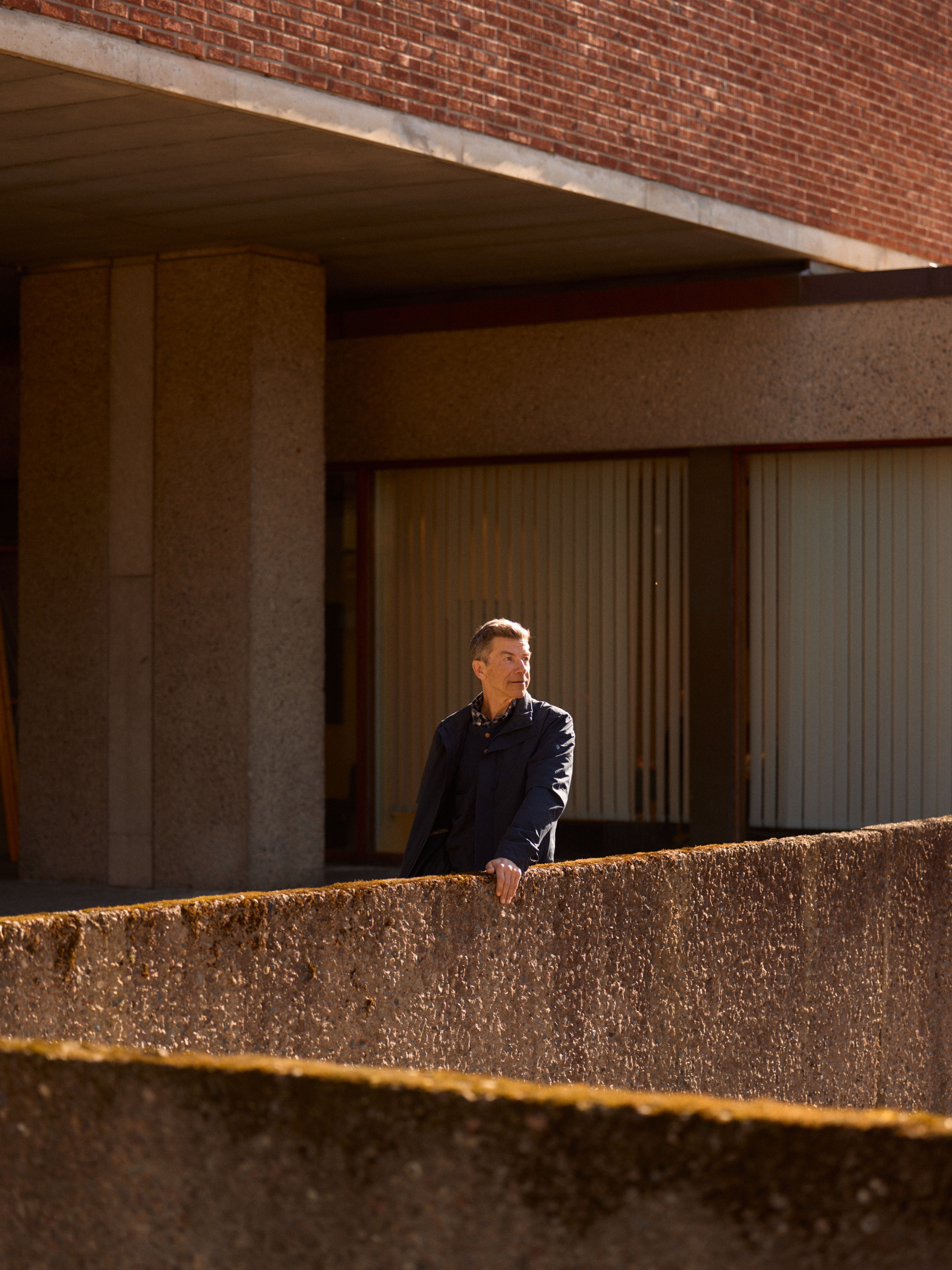 Mann i mørk jakke står på en terrasse foran en modernistisk bygning i betong og tegl, i sollys.