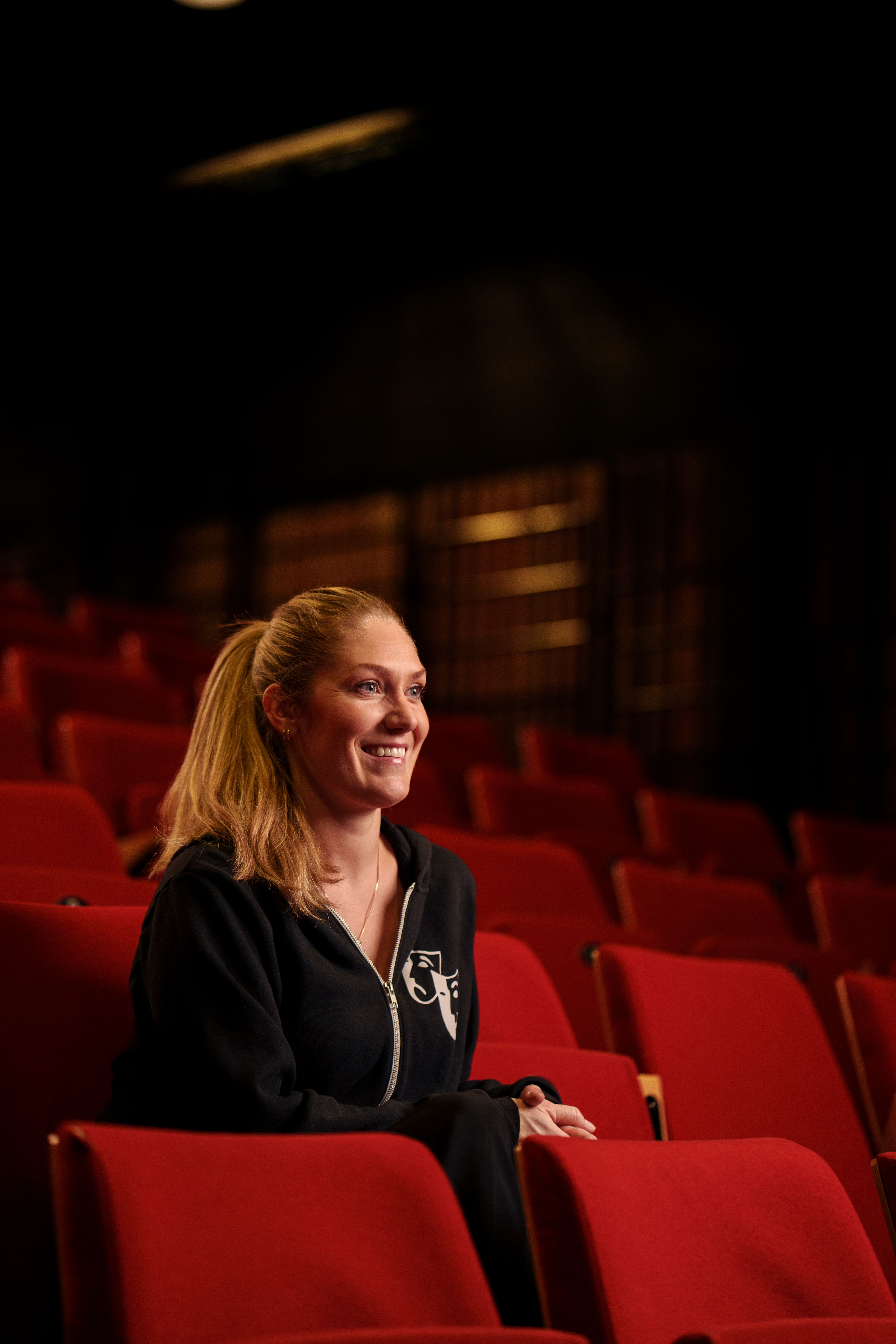 Mie Andberg, kledd i sort Action Teater-hettejakke, sitter i salongen og ser mot scenen med et smil.