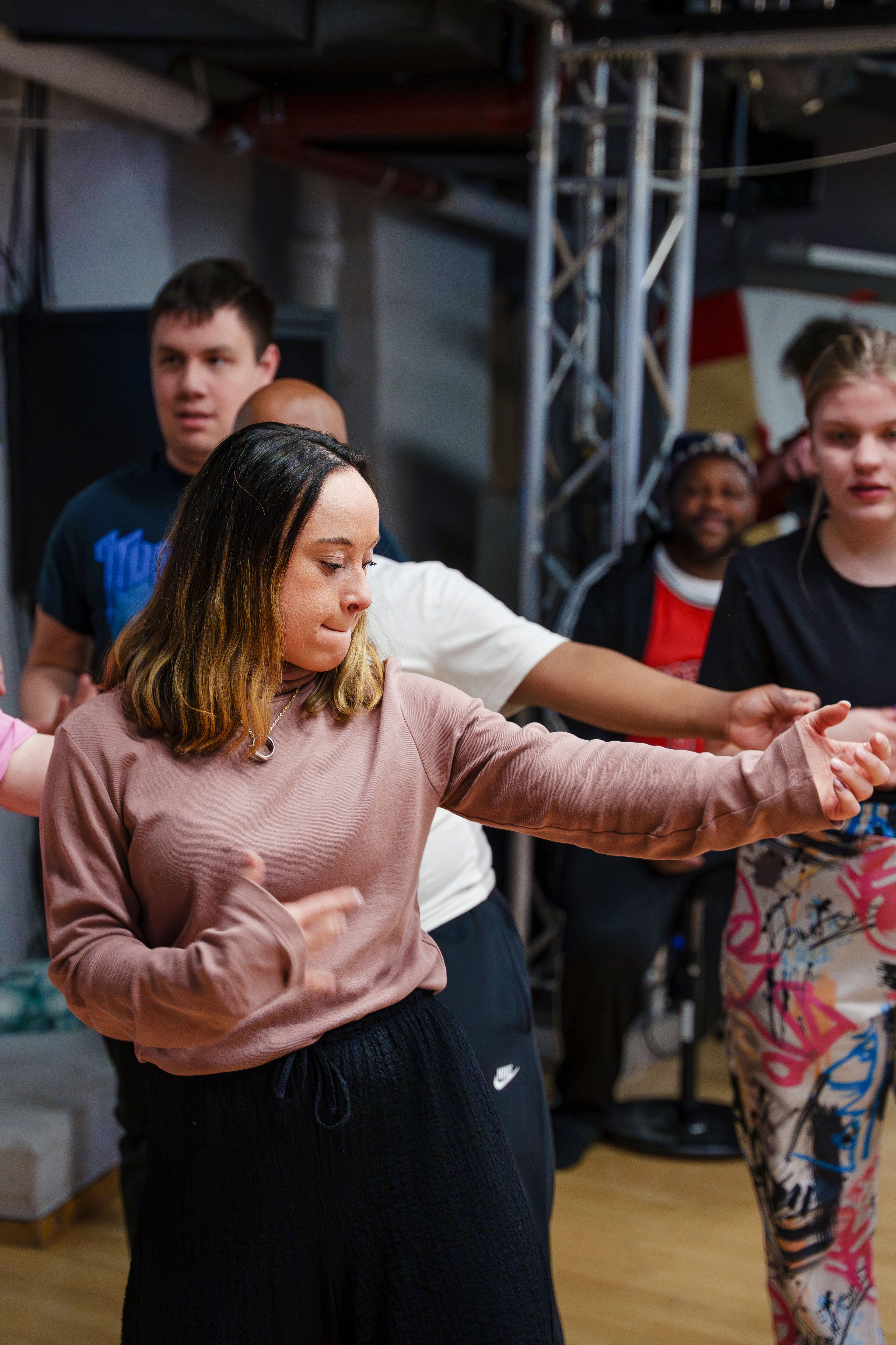 Kvinne i rosa genser danser med utstrakte armer mens andre deltakere er i bevegelse i bakgrunnen i ungdomshuset.
