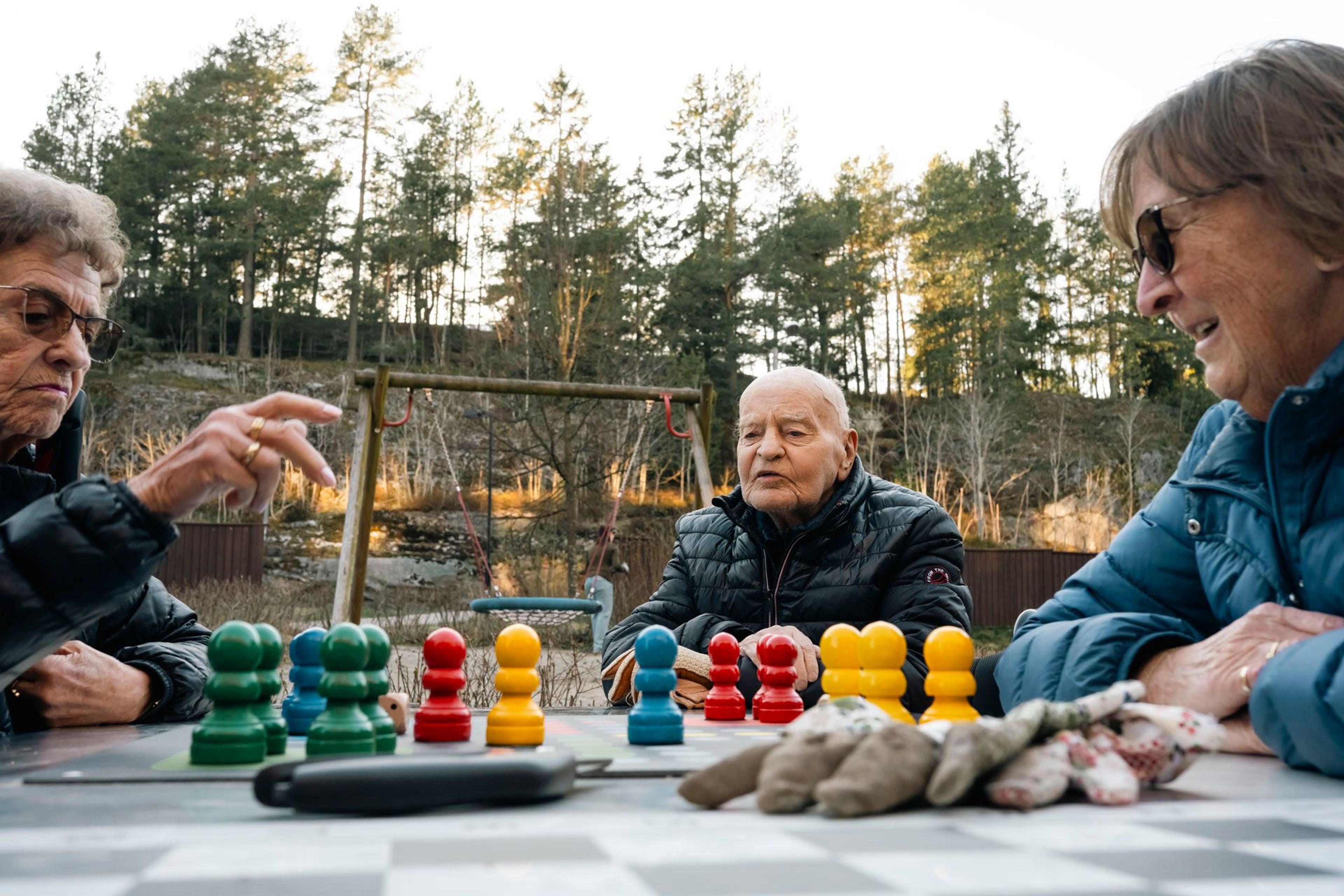 Tre eldre personer sitter rundt et utebord med fargerike ludobrikker og hagehansker, med husker bak.
