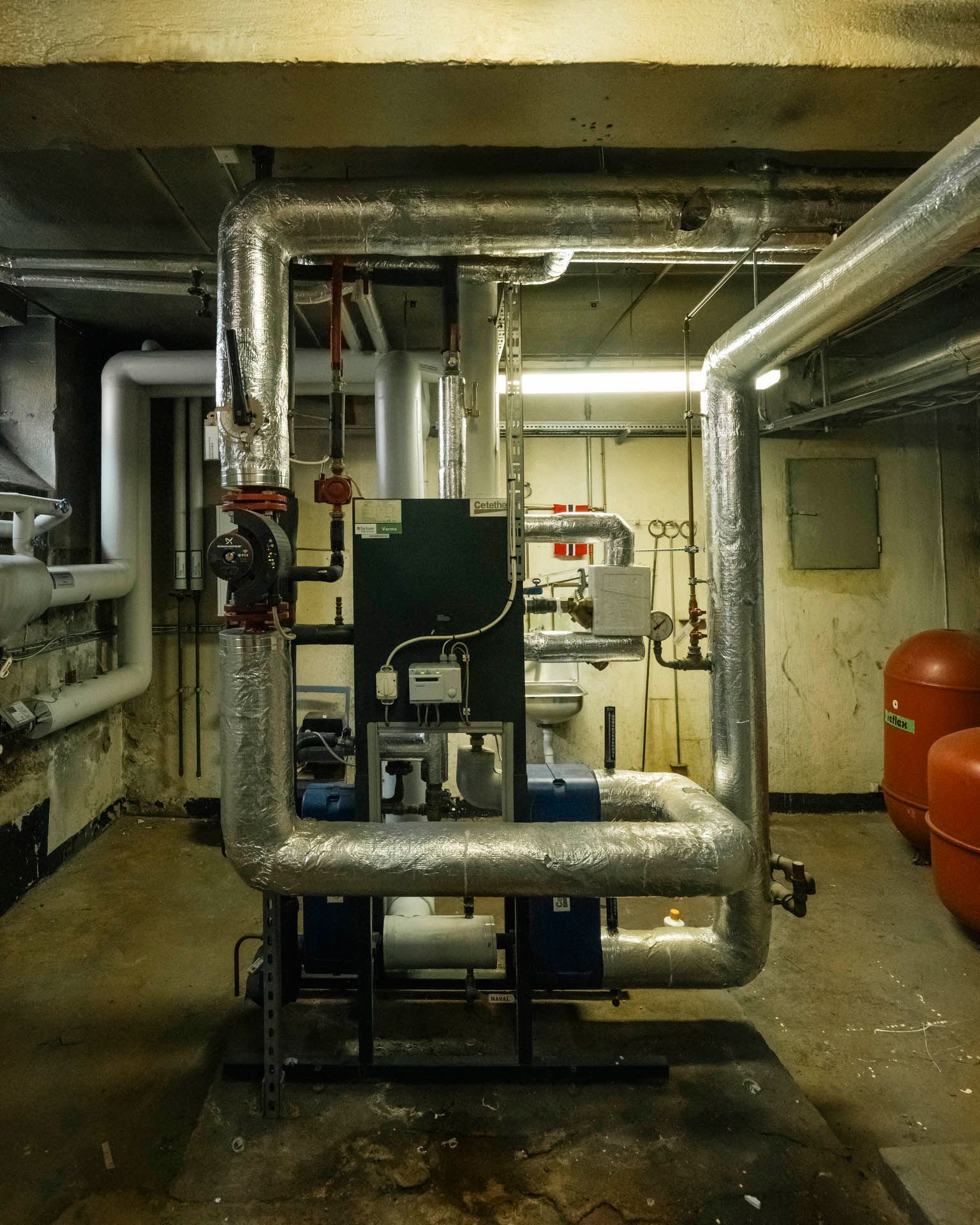 Et teknisk rom med store isolerte rør, pumper og utstyr som inngår i byggets varmeanlegg.