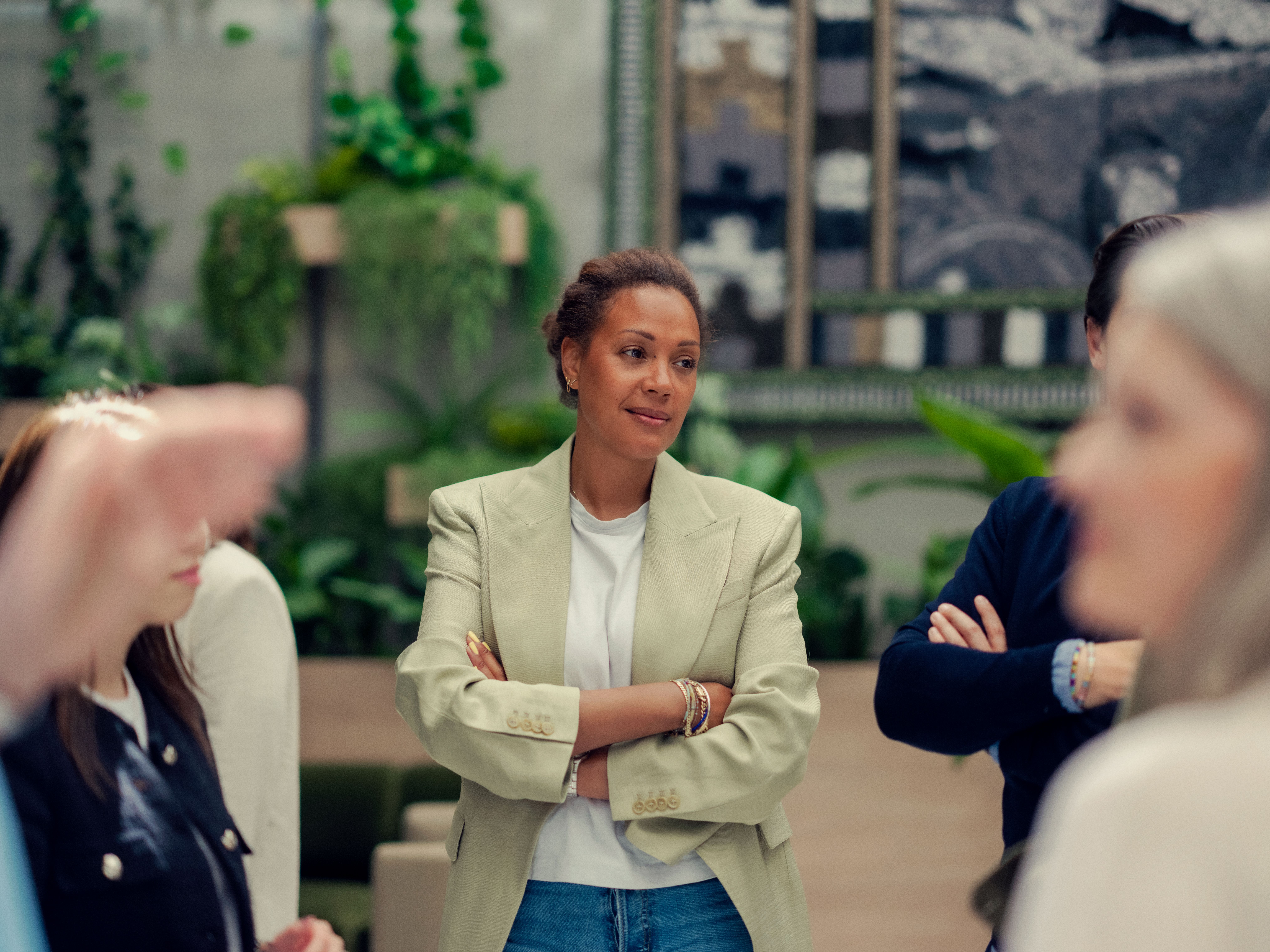 En kvinne i en lett blazer står med armene i kors og observerer oppmerksomt en diskusjon i et rom med planter og abstrakt kunst. Stemningen er tankefull.